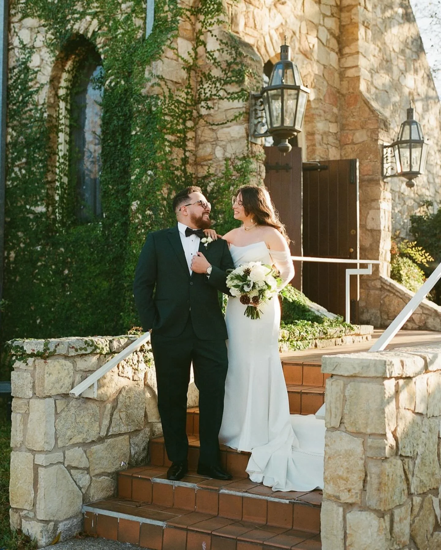 A bride and groom standing on stone steps outside a rustic building with stone walls and lanterns, smiling and looking at each other, during a wedding celebration.
