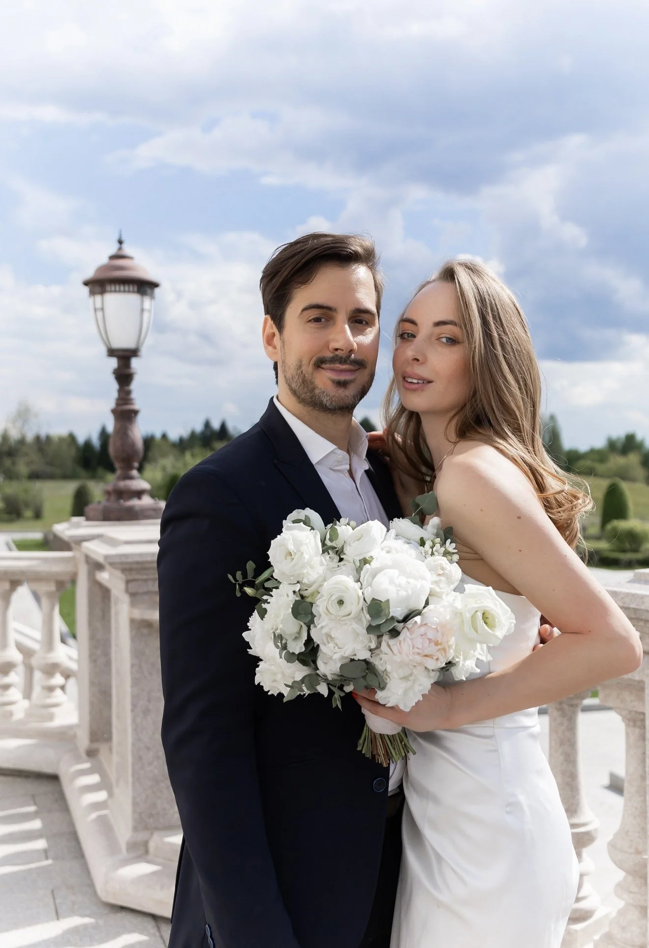 A newlywed couple posing outdoors on a stone balcony with a lamp post and green landscape in the background under a cloudy sky. The bride is holding a large bouquet of white flowers, and they are both smiling at the camera.