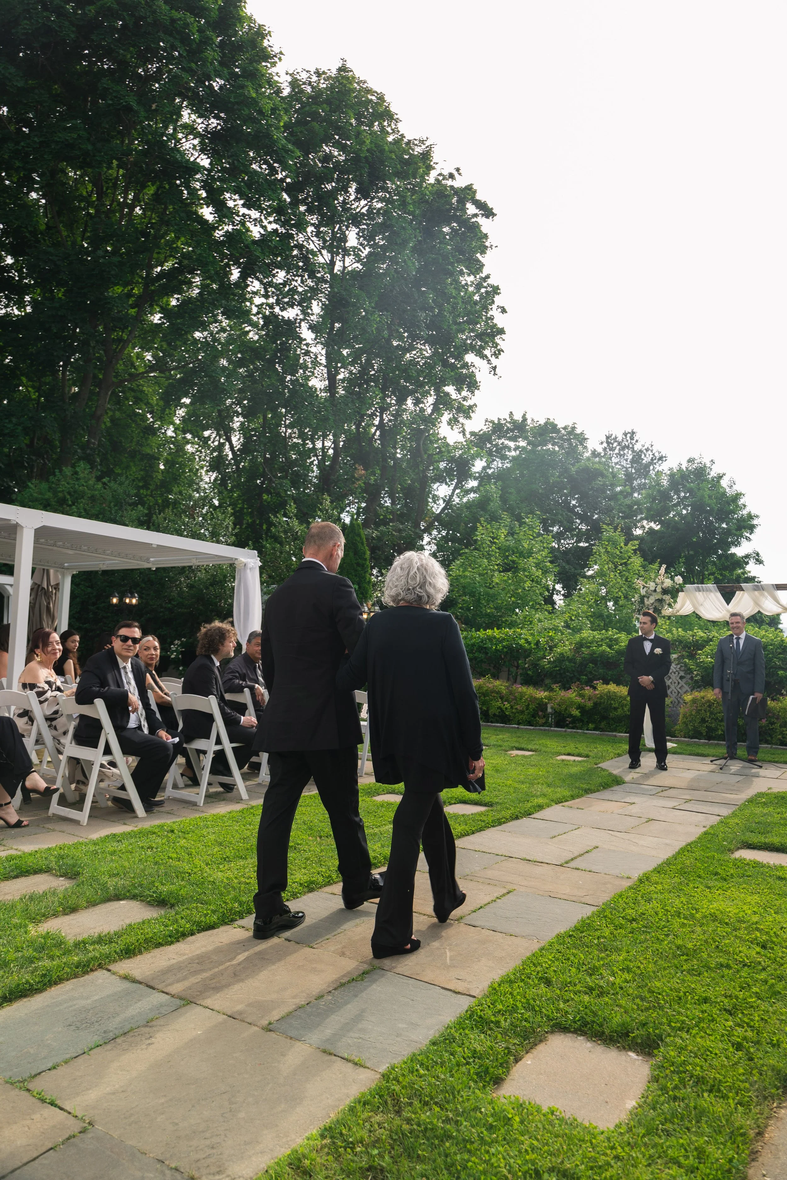 An outdoor wedding ceremony with guests seated on white chairs, a couple walking on a stone pathway, and a man and woman, possibly the officiant, walking toward the ceremony area with guests and greenery in the background.