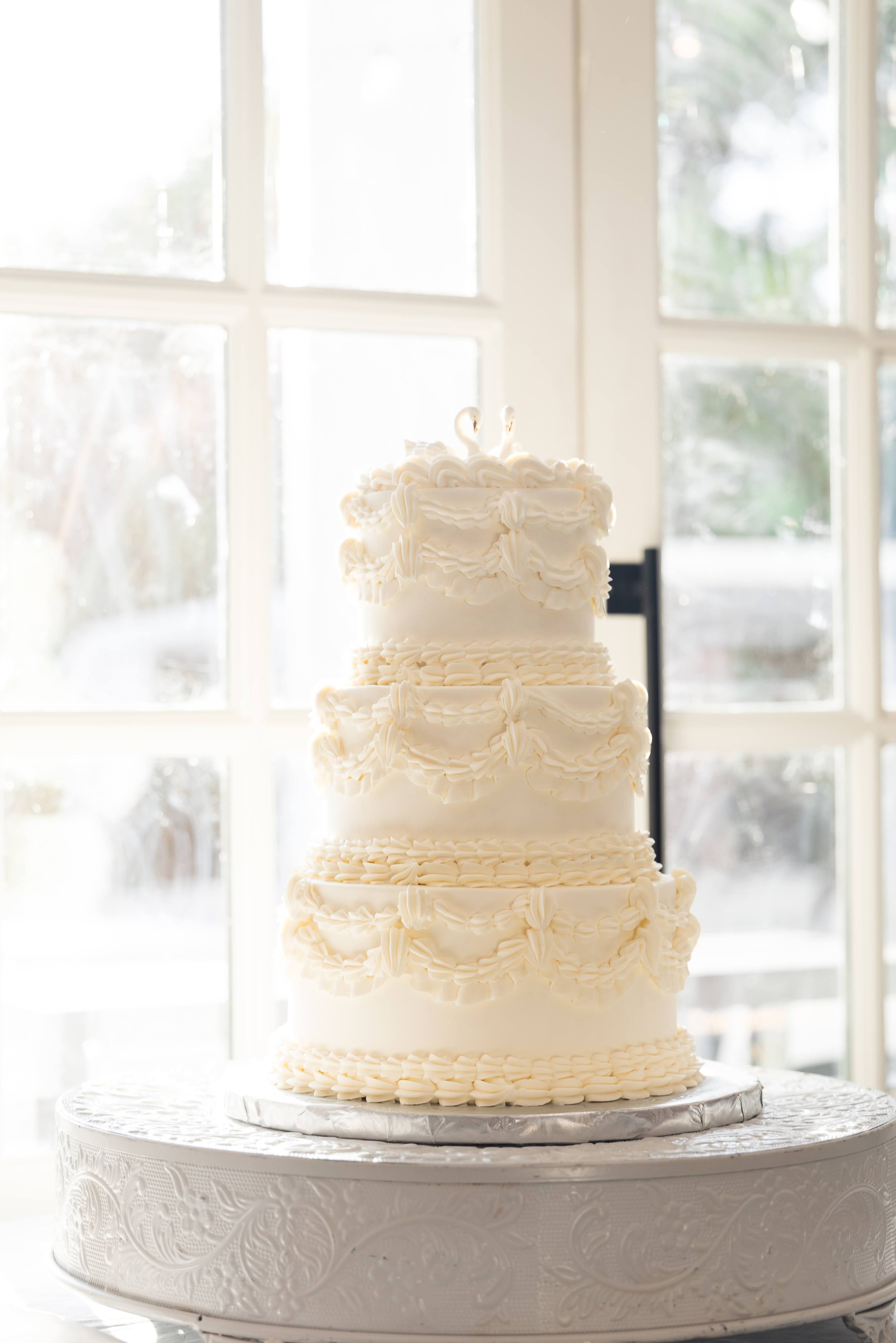A tall, multi-layered white wedding cake with decorative icing swirls, placed on a white lace-covered cake stand near a large window with daylight streaming in.