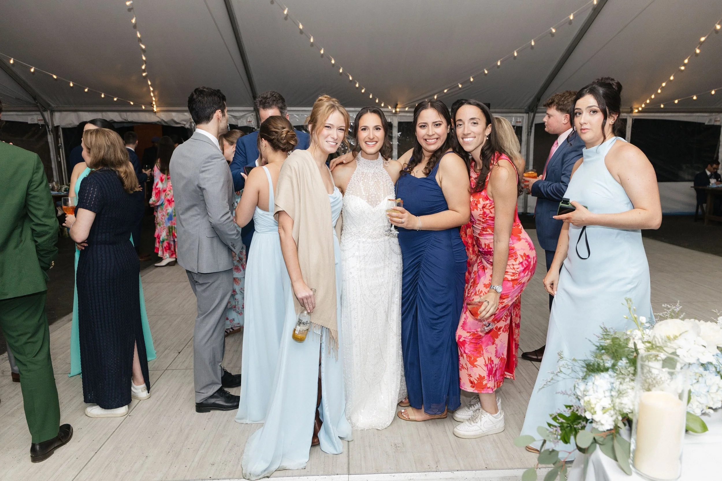 Group of women in formal dresses at a celebration or wedding under a large tent with string lights, smiling and posing for a photo.