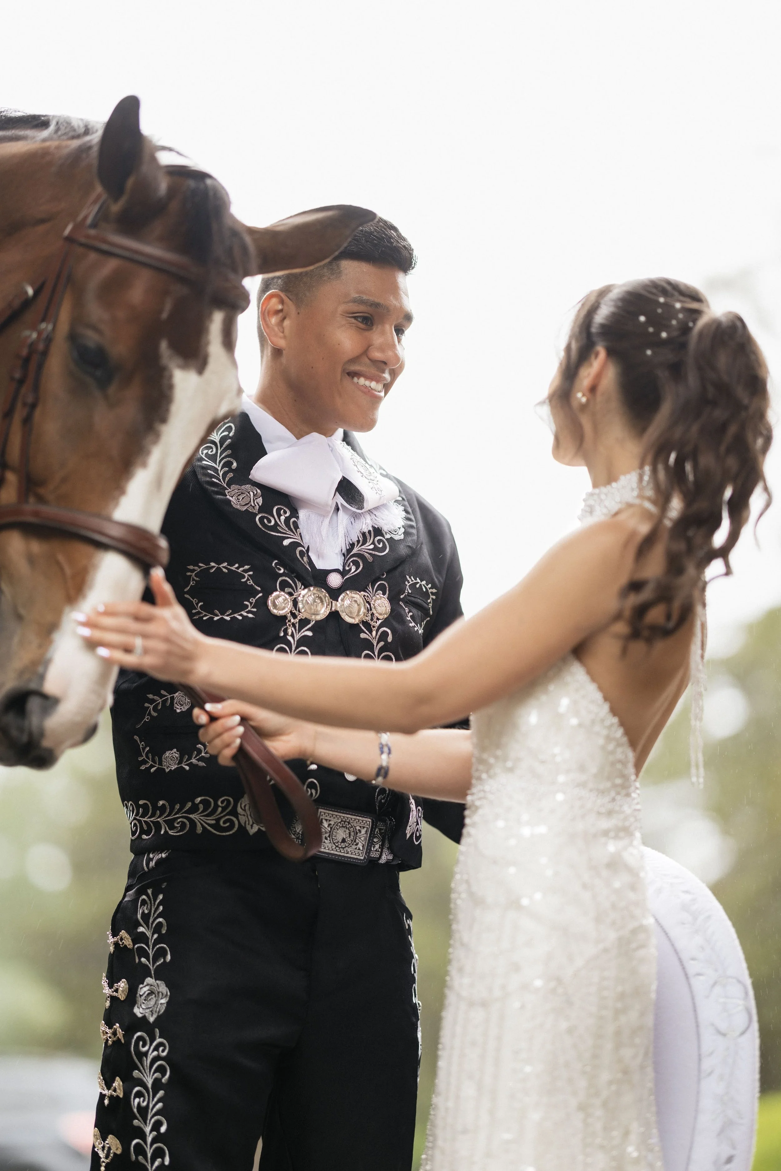 A bride and groom holding hands and looking at each other during their wedding ceremony outdoors, with a horse leaning over them.