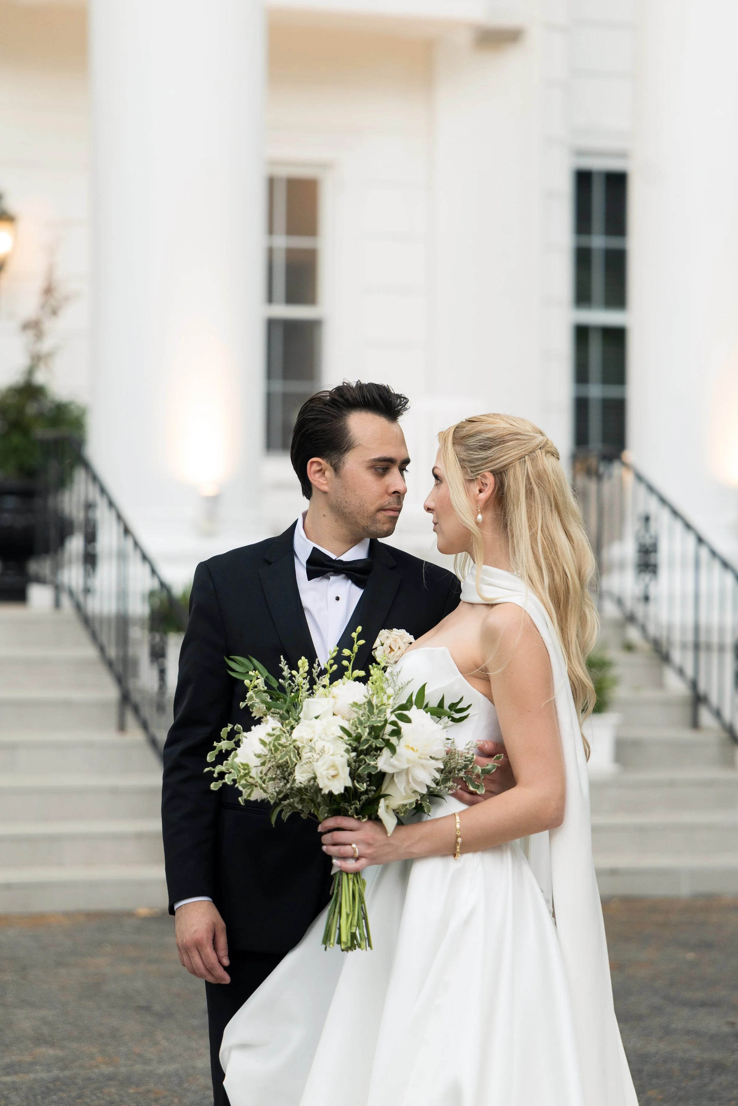 A bride and groom are standing close together outside a white building with columns, looking into each other's eyes. The bride is holding a large bouquet of white flowers and has long blonde hair styled in loose waves, wearing a white wedding dress. 