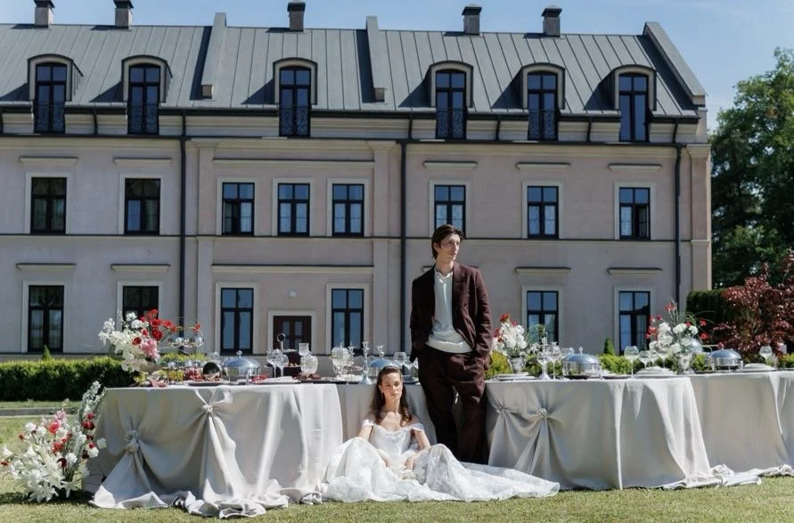A young girl in a white dress sitting on the ground, and a young man in a brown suit standing behind a long table with a white tablecloth, set outdoors in front of a large, multi-story building with multiple windows and a blue roof, during daytime wi