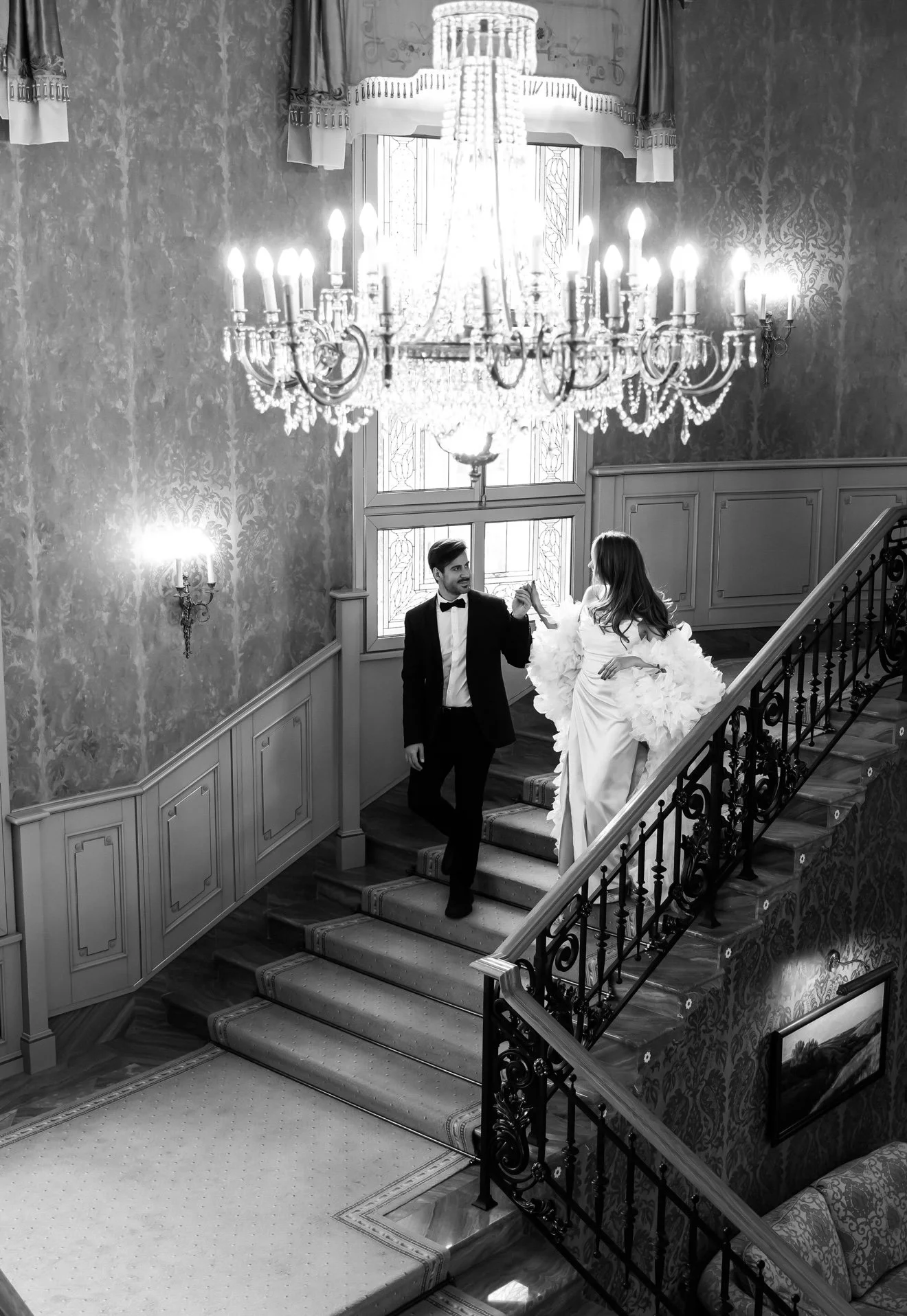 A black and white photo of a formal couple descending a staircase in an opulent interior. The man is in a tuxedo and the woman is in a wedding dress with large floral details. A chandelier hangs above them, and decorative wallpaper covers the walls. 
