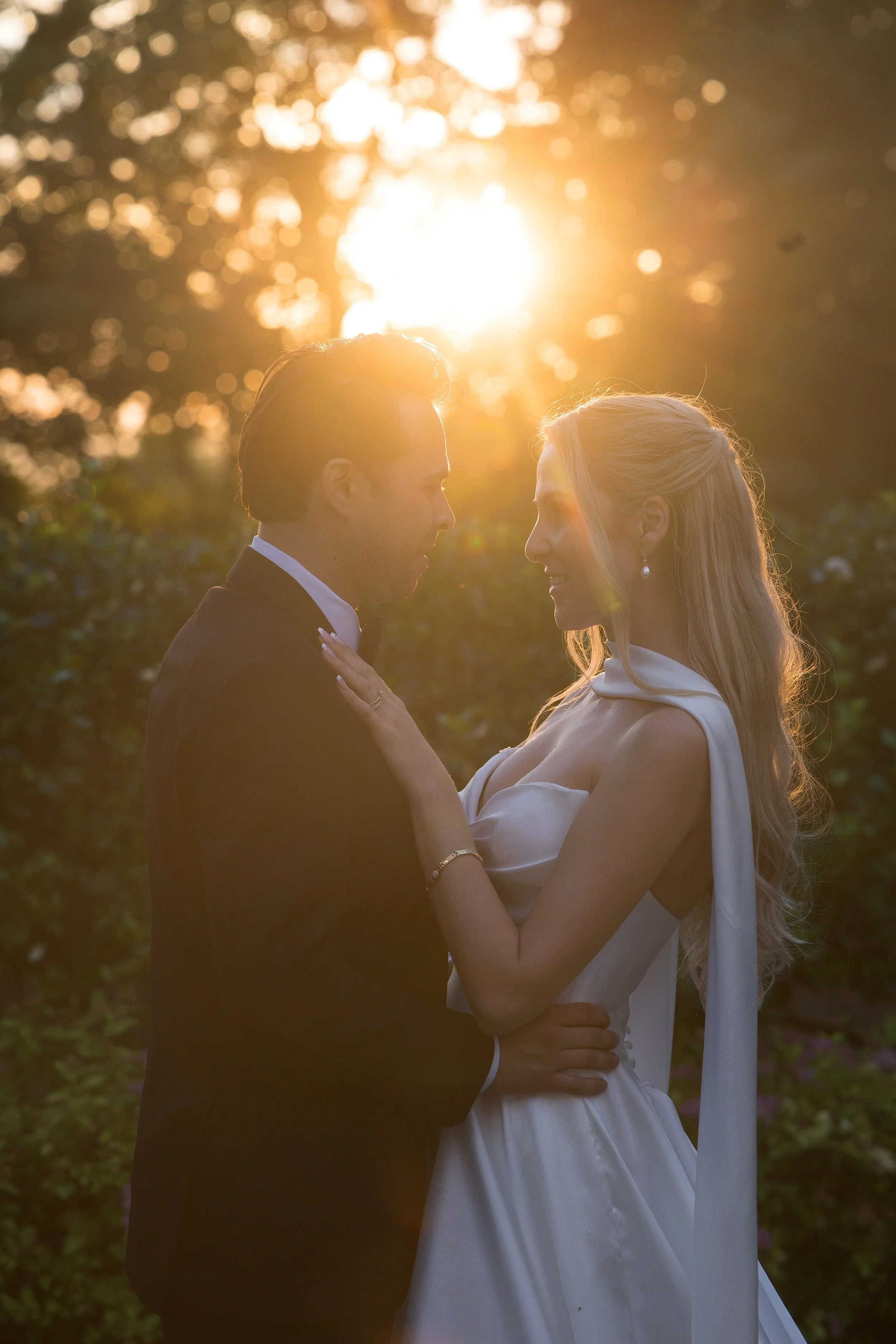 A couple in wedding attire standing close together outdoors during sunset, with the sun shining through trees in the background.