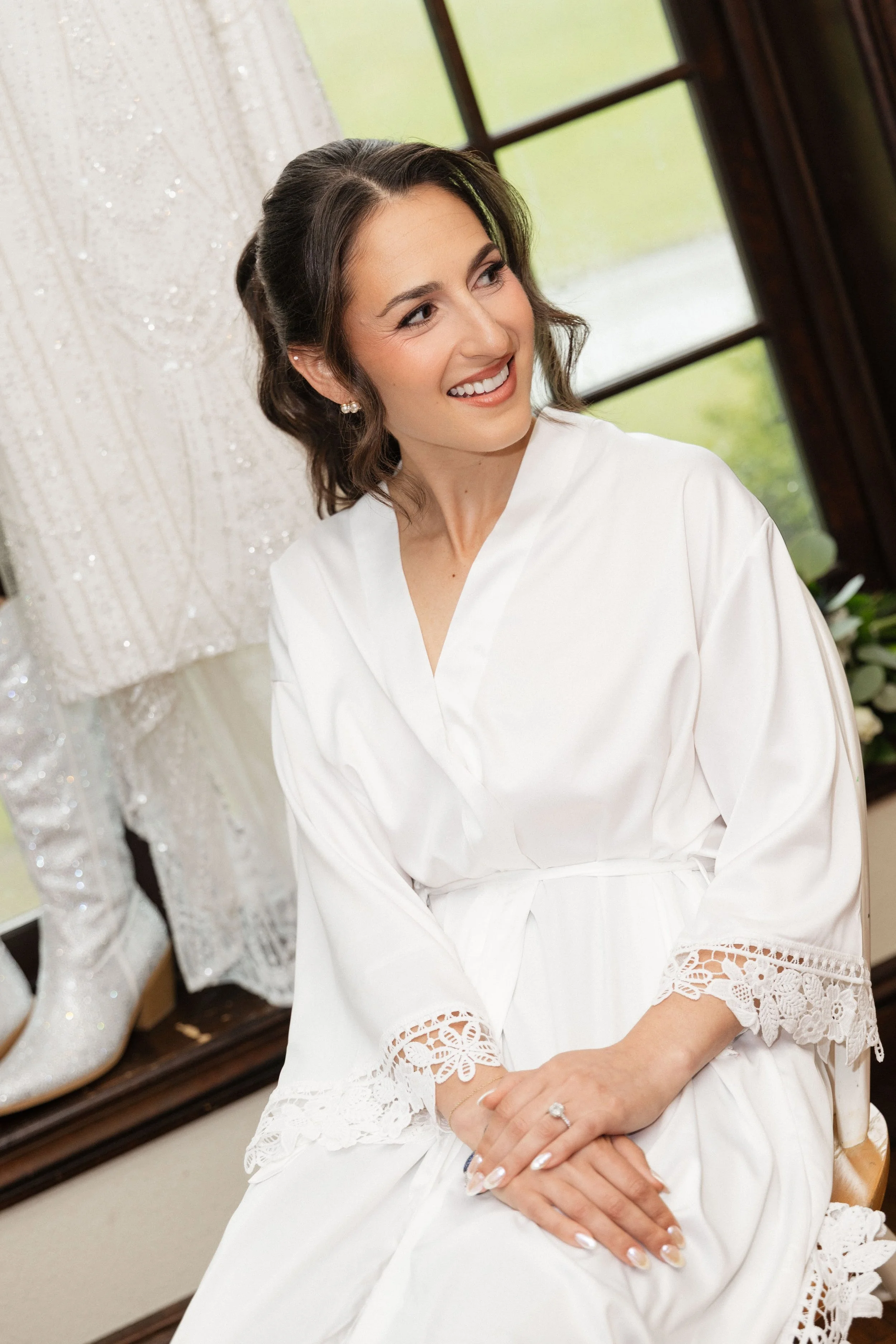 A woman smiling in a white robe, sitting beside a display of white lace and beaded shoes.
