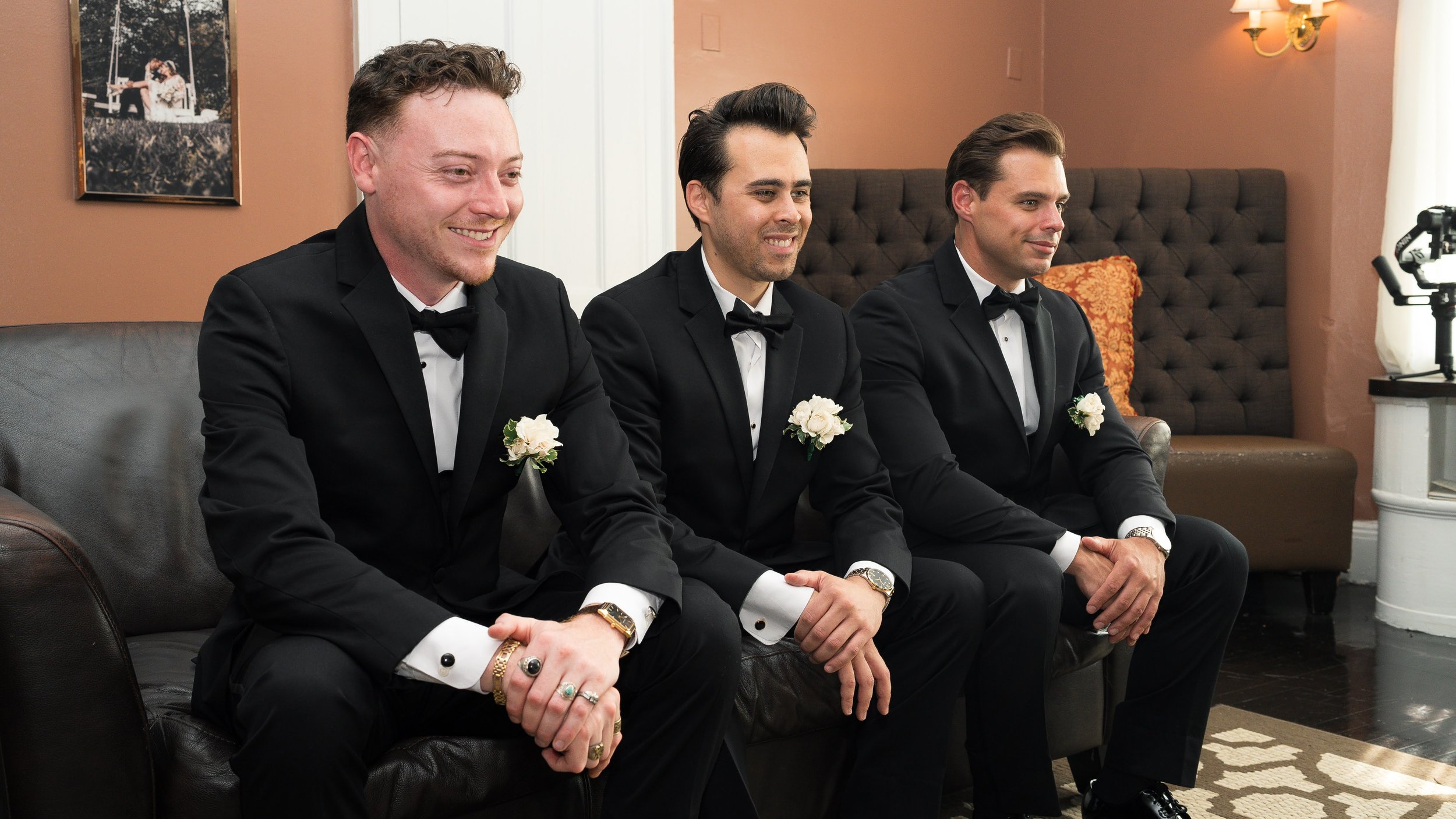 Three men dressed in black tuxedos with white shirts and black bow ties, sitting on a leather couch during a formal event, each wearing floral boutonnières and jewelry.