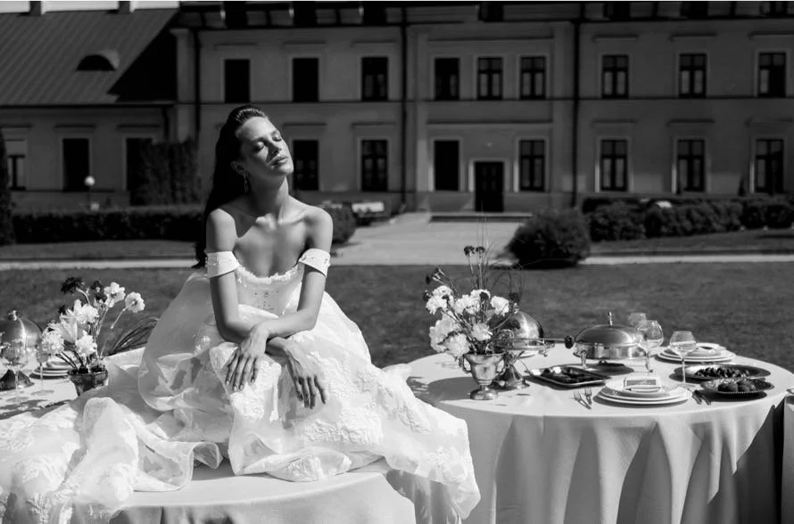 A woman in a wedding dress sitting on an outdoor banquet table set with plates, flowers, and serving dishes in front of a large building.