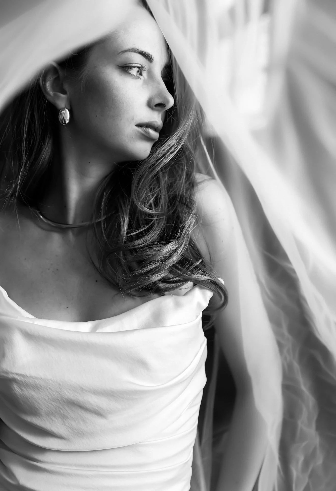 A black and white photograph of a woman with wavy hair, wearing earrings and a necklace. She is looking to her right with a calm expression, and is wearing a light-colored off-shoulder top.