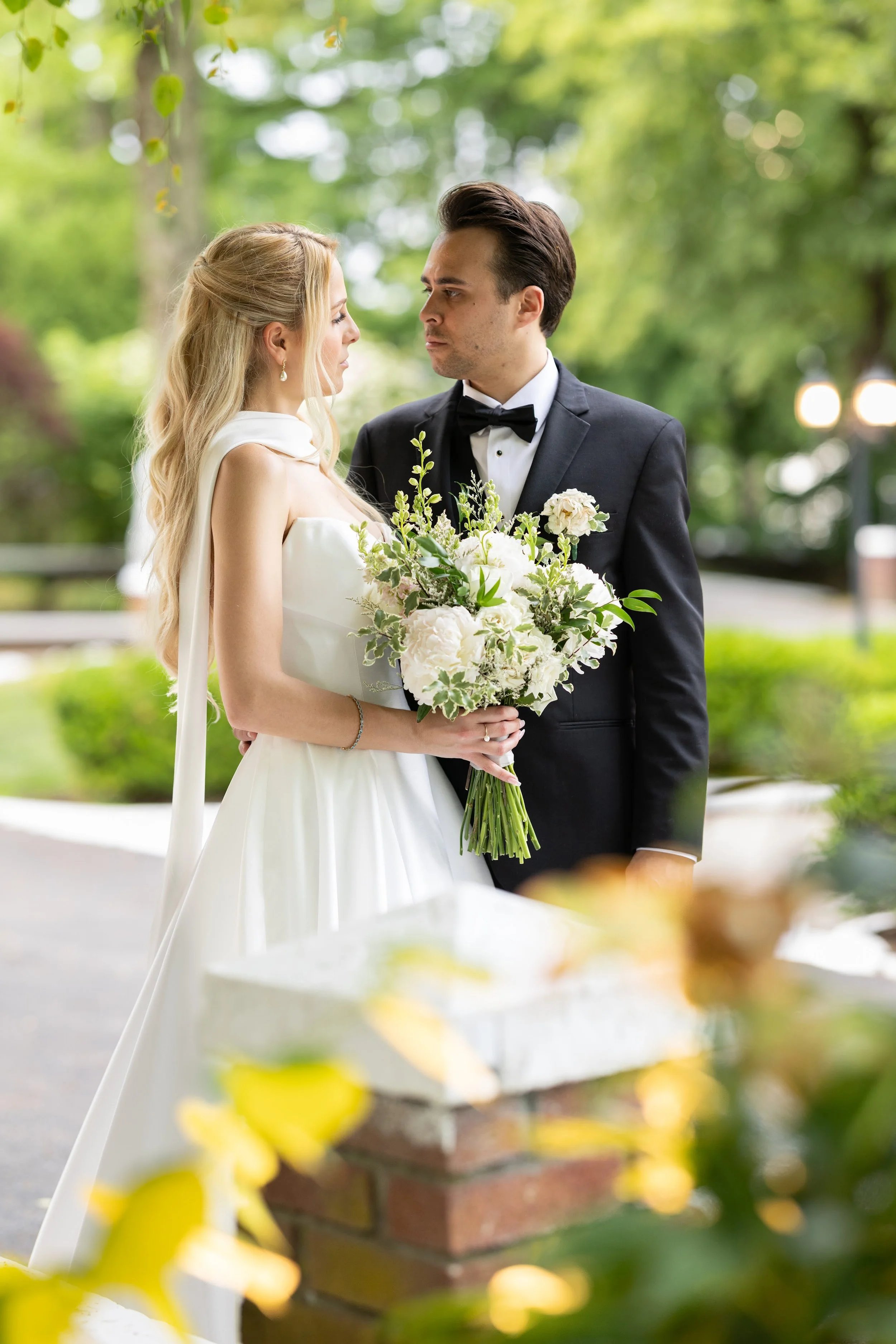A bride and groom face each other outdoors, with the bride holding a bouquet of white and green flowers. The bride is wearing a white wedding dress and pearl earrings, while the groom is in a black tuxedo with a black bow tie. The background is a lus