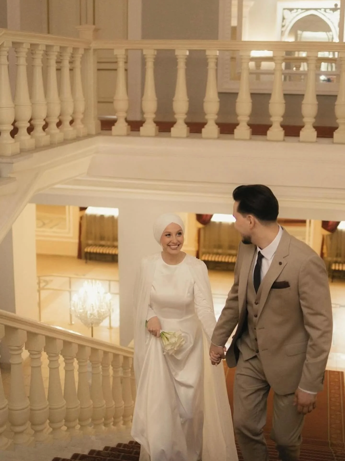 A bride and groom holding hands and walking upstairs in a luxurious, ornate hotel or mansion with warm lighting and a chandelier in the background.
