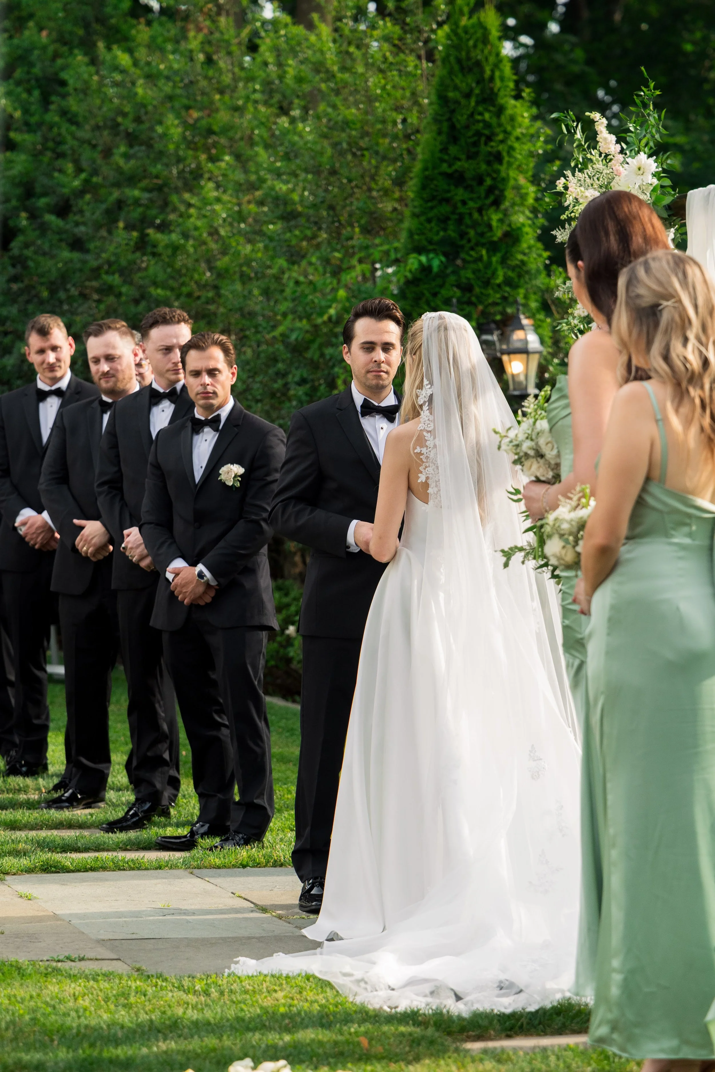 A wedding ceremony outdoors with a group of men in tuxedos and women in pastel dresses, including a bride in a white dress and veil, holding flowers. The bride and groom are facing each other, standing on a grassy area with trees and decorative flowe