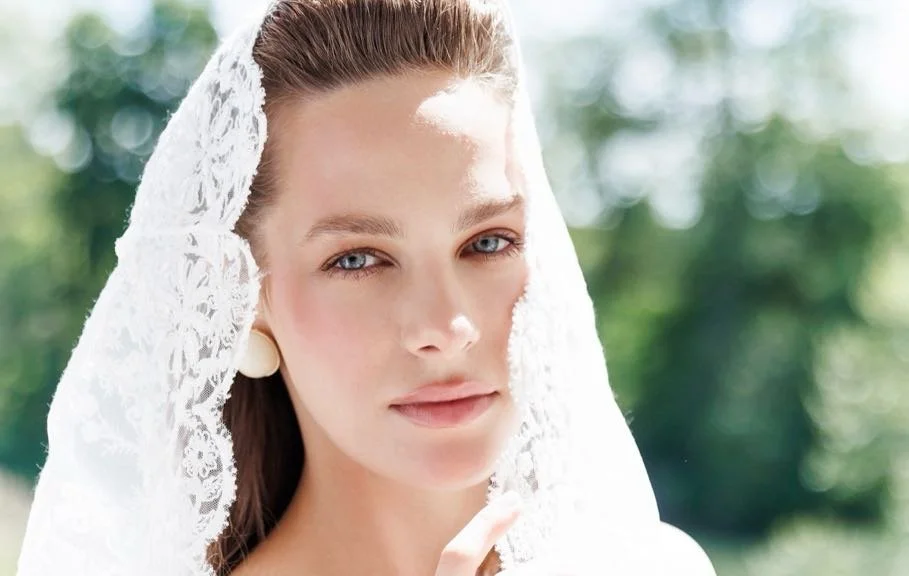 A woman with fair skin and blue eyes wearing a white lace head cover, outdoors with blurred green trees in the background.