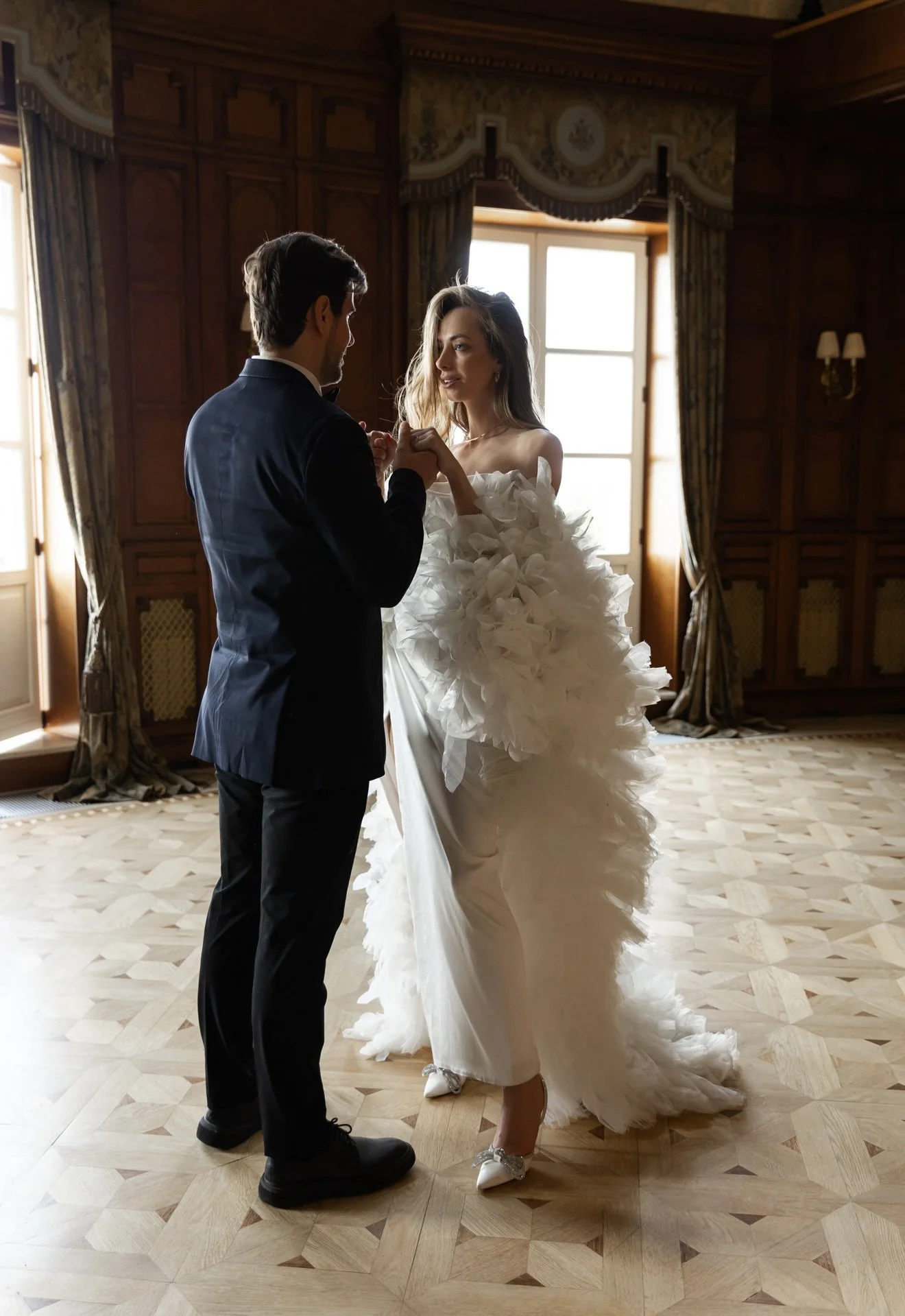 A bride and groom holding hands and dancing in a lavishly decorated room with wood-paneled walls and large windows, during their wedding celebration.
