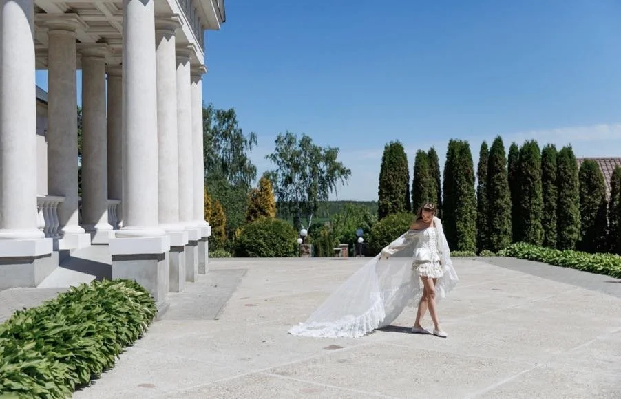 Bride in a white wedding dress with a long train standing outdoors near a classical building with columns and tall trees.