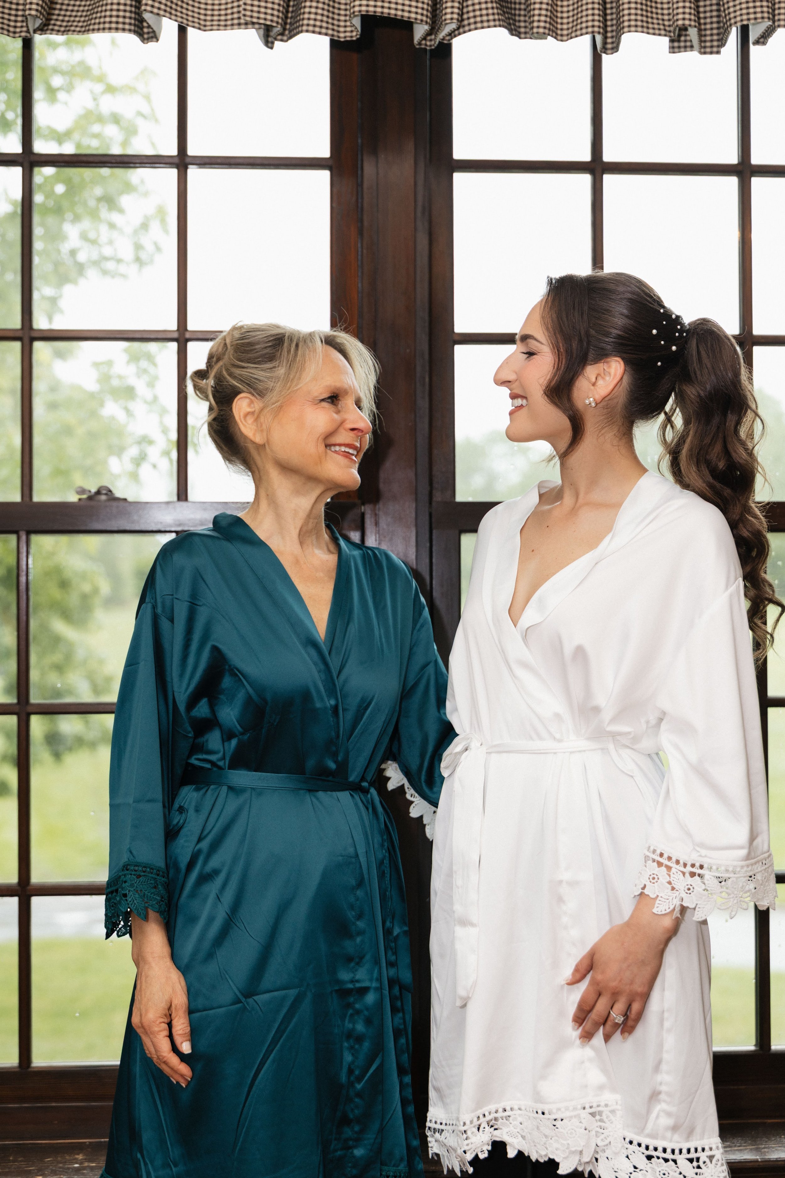 A bride in a white robe smiling at an older woman in a teal silk robe, both standing by a window in a cozy room.