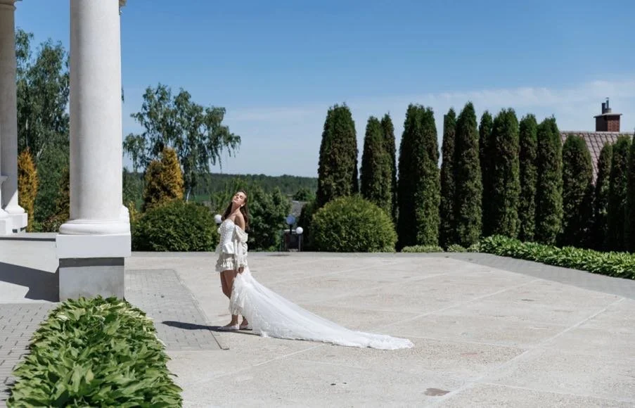 A woman in a white wedding dress standing outside on a sunny day, with a long train trailing behind her, near a building with columns and lush green trees and bushes in the background.