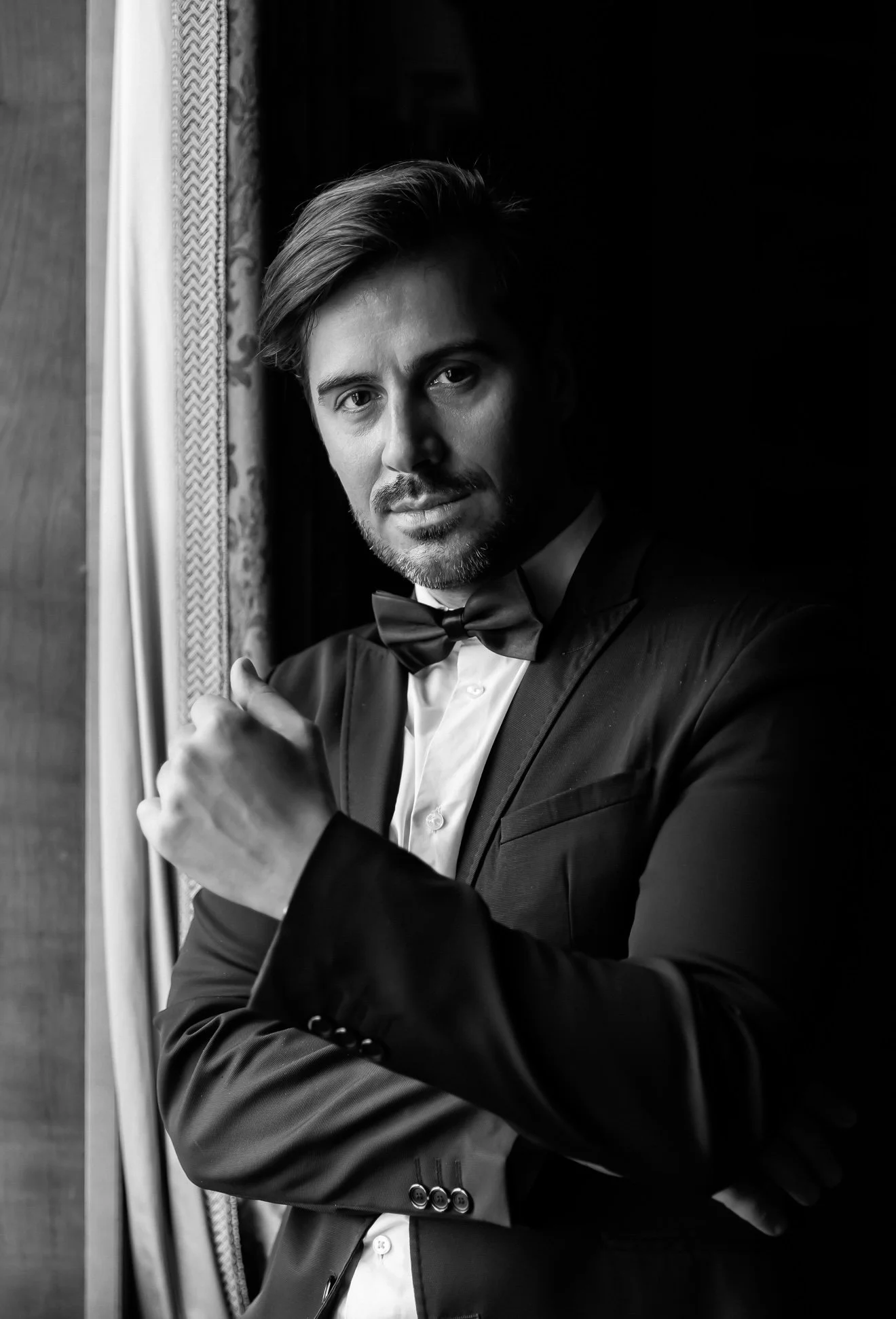 A well-dressed man in a tuxedo, with a bow tie, looking at the camera, standing indoors near a window with curtains, in black and white.