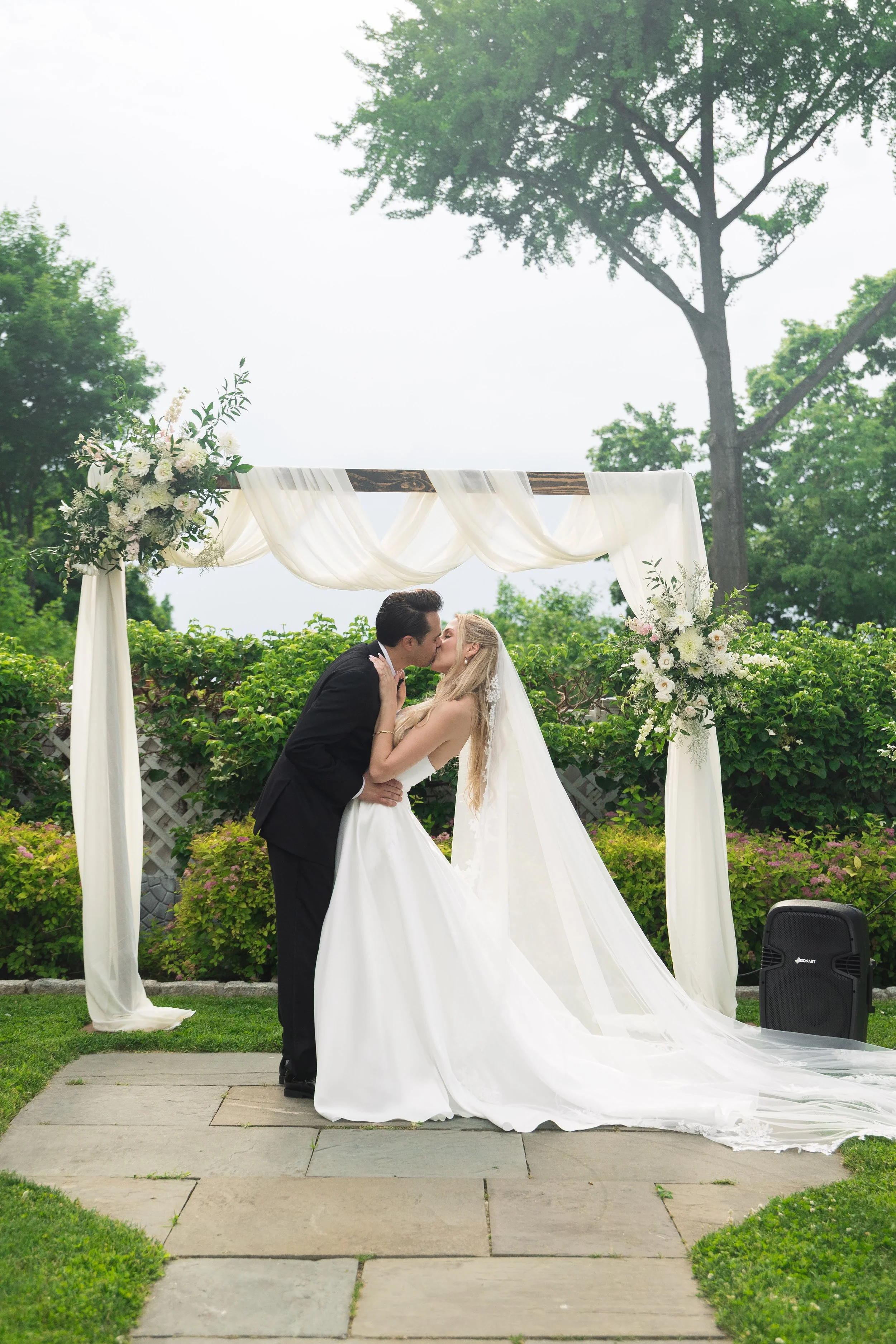 A bride and groom kiss under a decorated wedding arch outdoors surrounded by greenery.