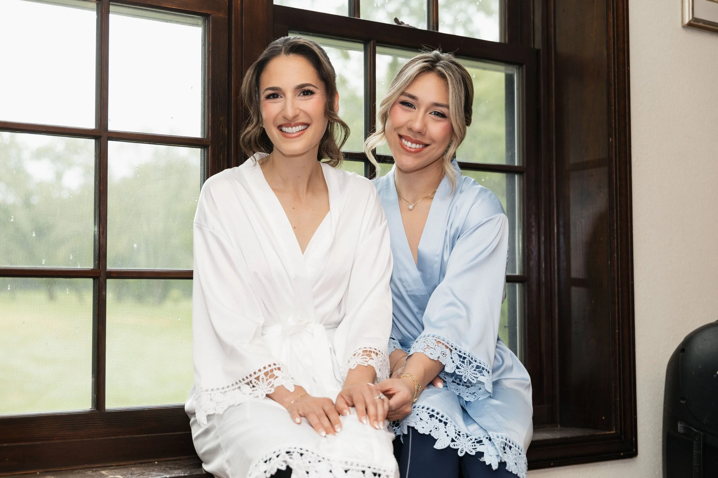 Two women smiling sitting closely in front of a large window with a wooded view, wearing light-colored and pastel tops with lace details.