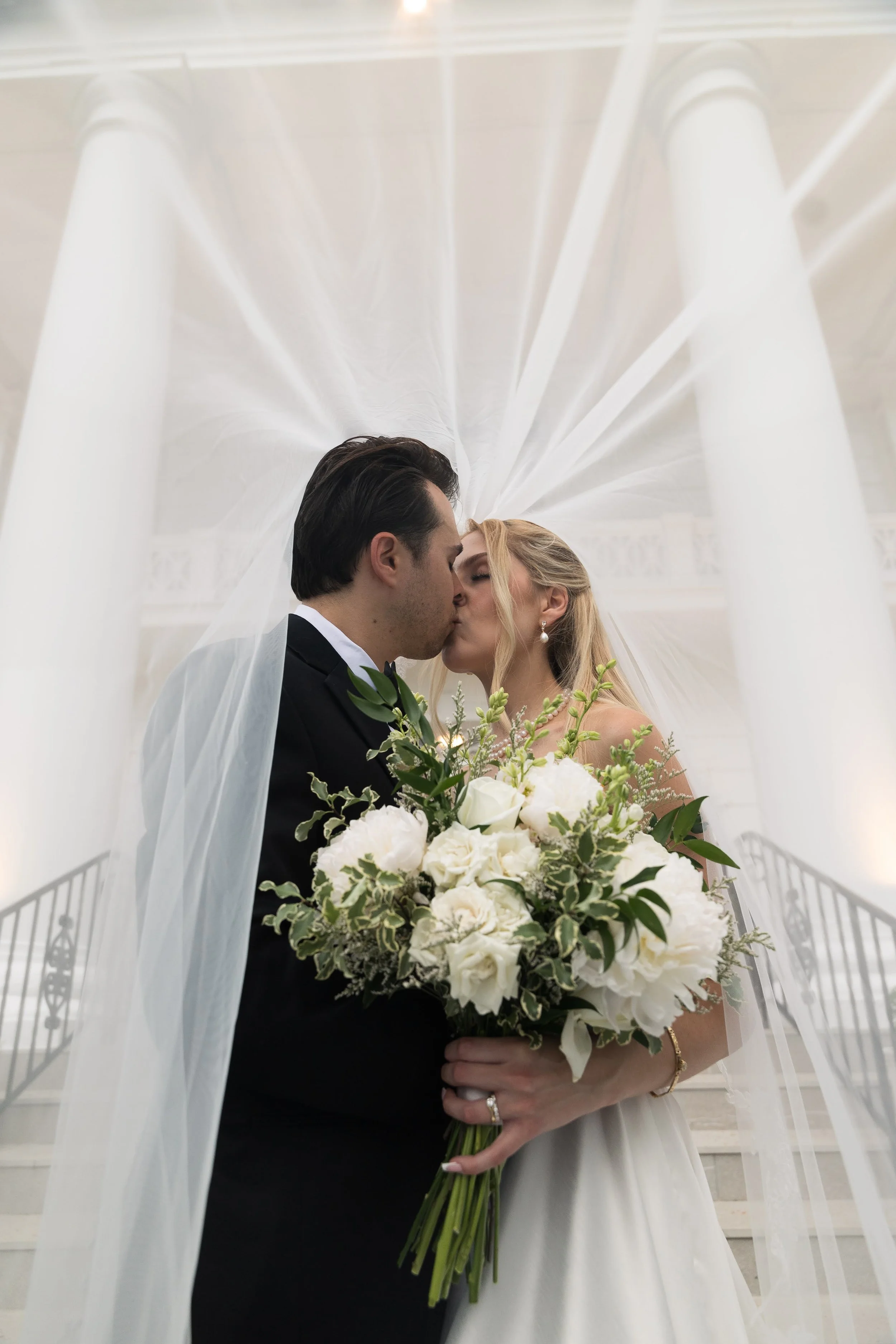 A bride and groom sharing a kiss underneath a wedding veil in a white, elegant church setting, with the bride holding a large bouquet of white flowers and greenery.