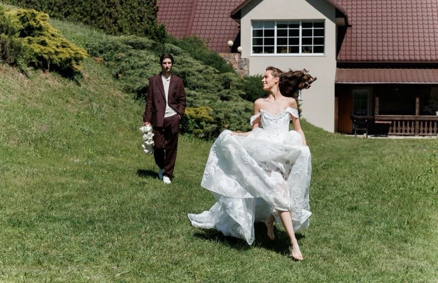 Bride in a white wedding dress running on grass, groom in a dark suit walking behind, house with a red roof in the background.