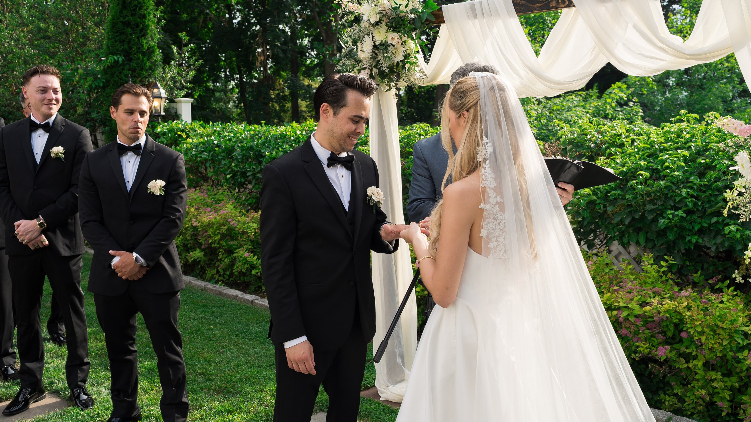 A bride and groom exchanging vows at an outdoor wedding ceremony, with groomsmen standing in the background, under a decorated arch with greenery and white flowers, in a garden setting.
