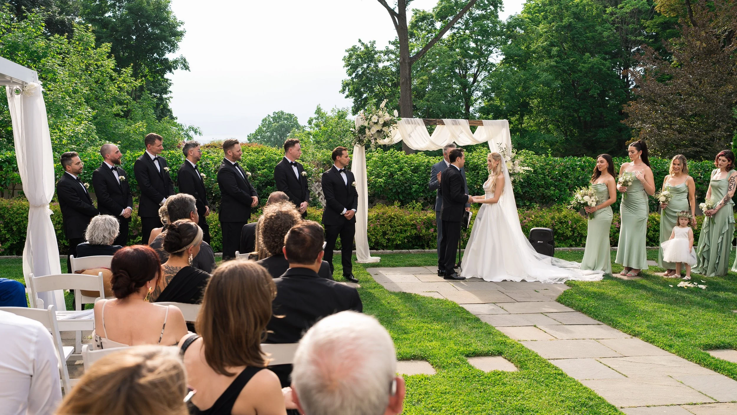 A wedding ceremony taking place outdoors on a lawn, with a bride and groom exchanging vows under a decorated arch. Bridesmaids in green dresses and groomsmen in black tuxedos stand to the sides, while guests sit in white chairs watching the event.