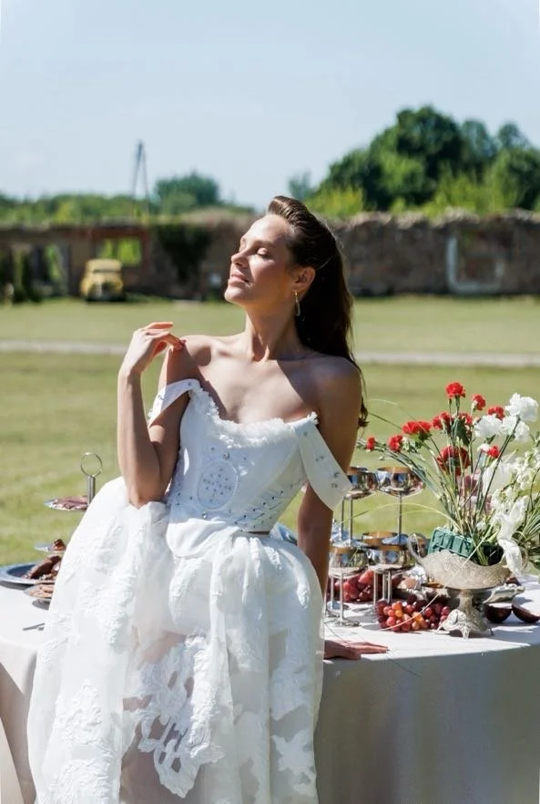 Woman in a white dress, enjoying the outdoors at a table with flowers, food, and drinks on a sunny day.
