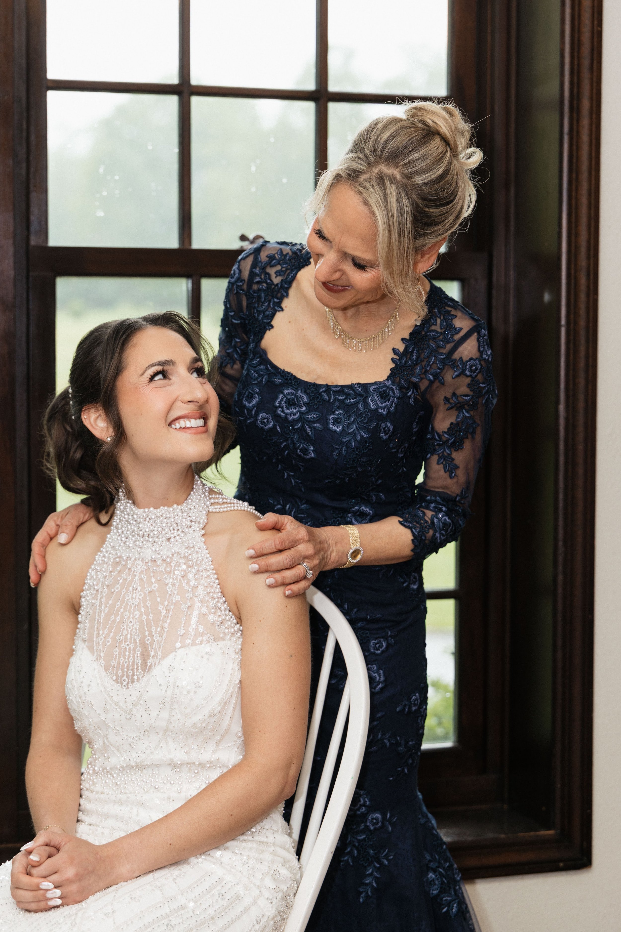 A smiling young woman in an elegant white beaded dress sitting on a white chair, looking up at an older woman in a dark blue lace dress who is standing beside her, smiling and looking down at her. They are indoors near a large window with a wooden fr