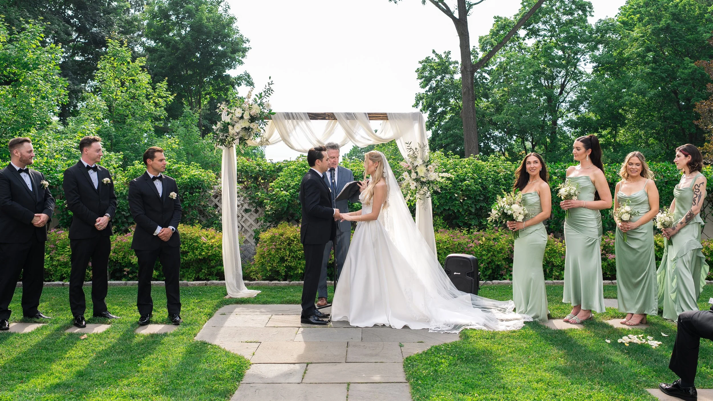 A wedding ceremony outdoors with the bride and groom exchanging vows under a decorated arch, surrounded by bridesmaids and groomsmen in formal attire, with a lush green garden in the background.