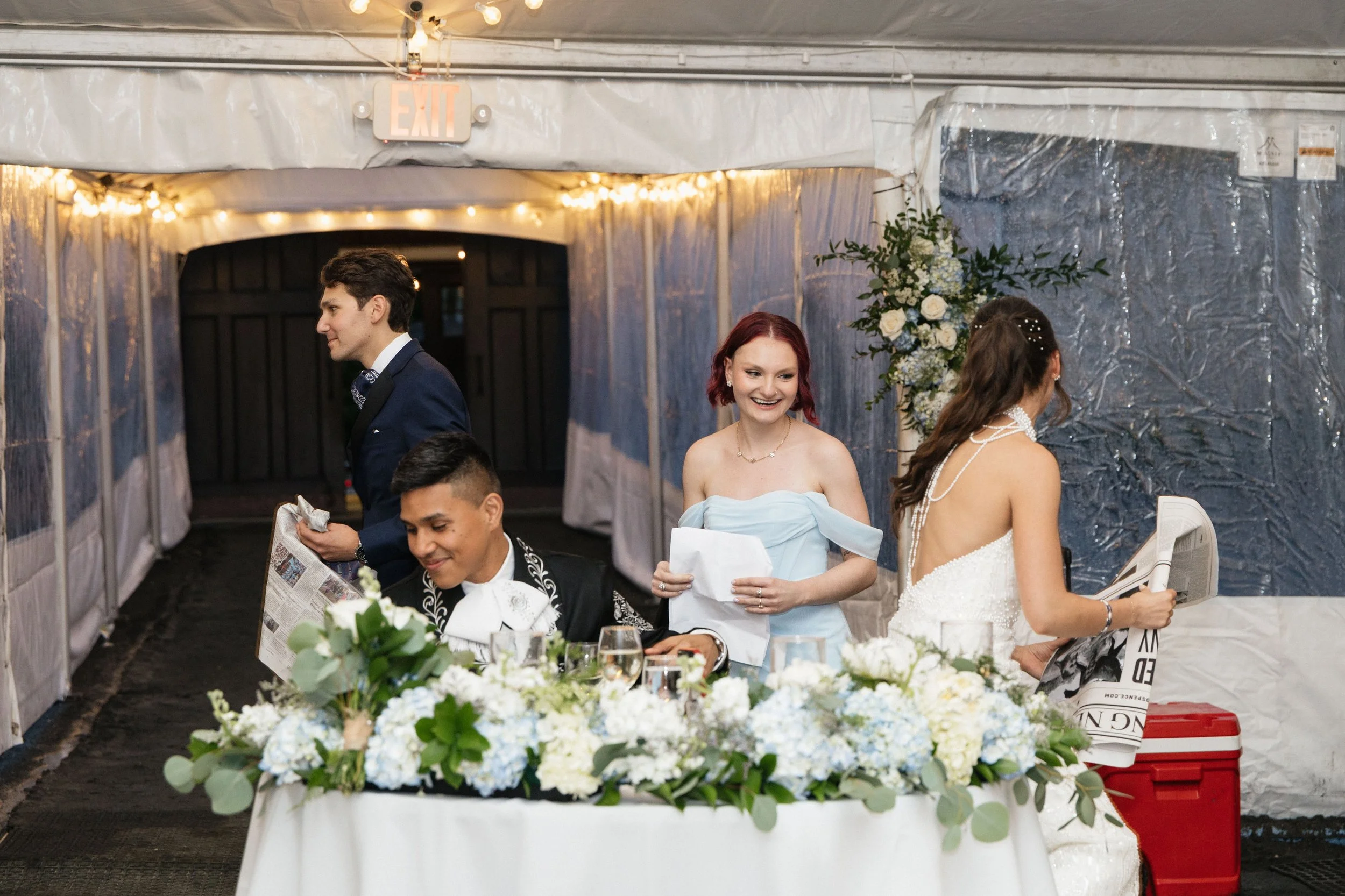 People at a wedding reception, with a woman in a white dress and red hair winking and smiling, a man in a tuxedo, and other guests sitting at a table decorated with white flowers and greenery inside a tent.