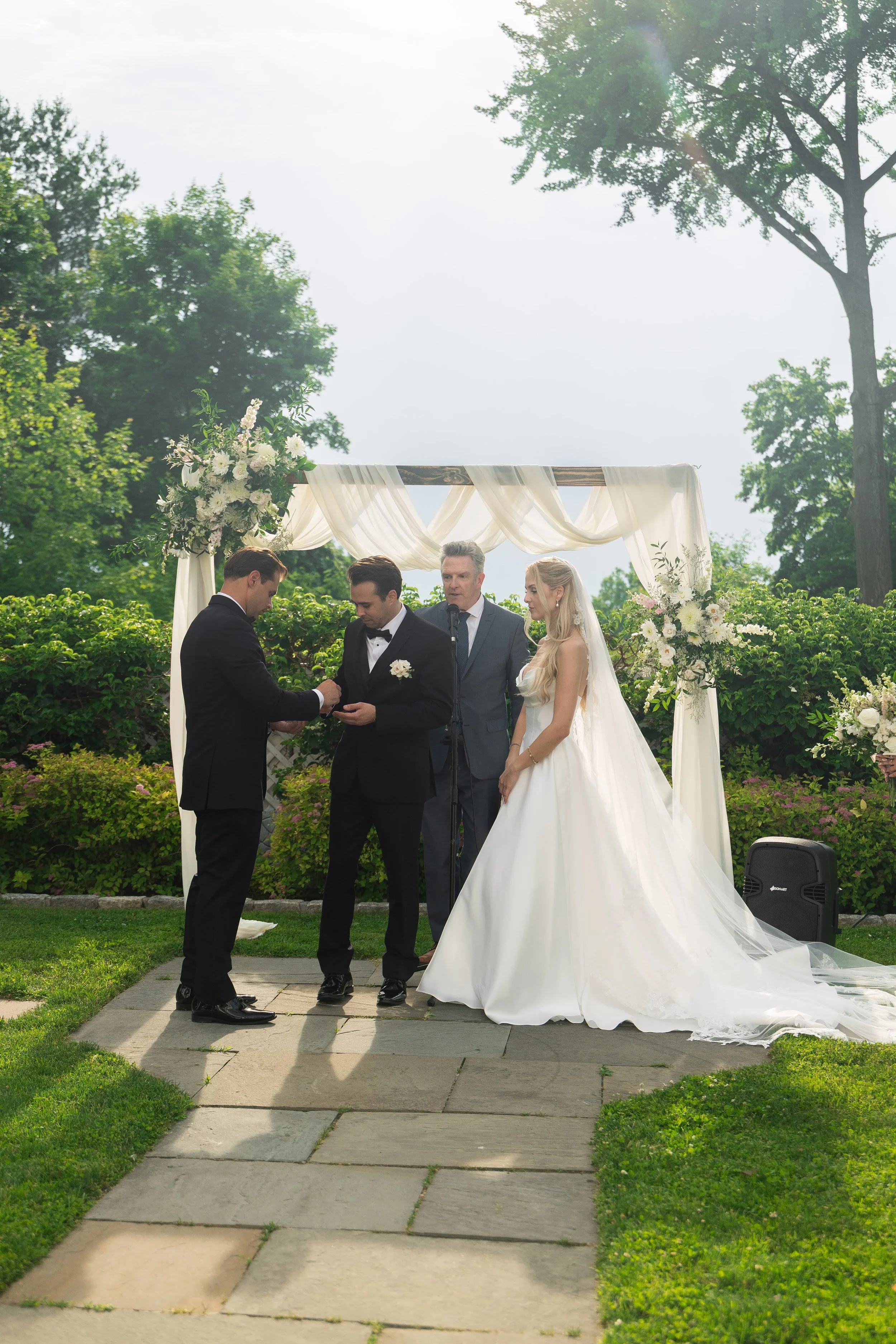 A couple and an officiant in a wedding ceremony outdoors under a decorated arch with white flowers and fabric, surrounded by greenery, during daylight.