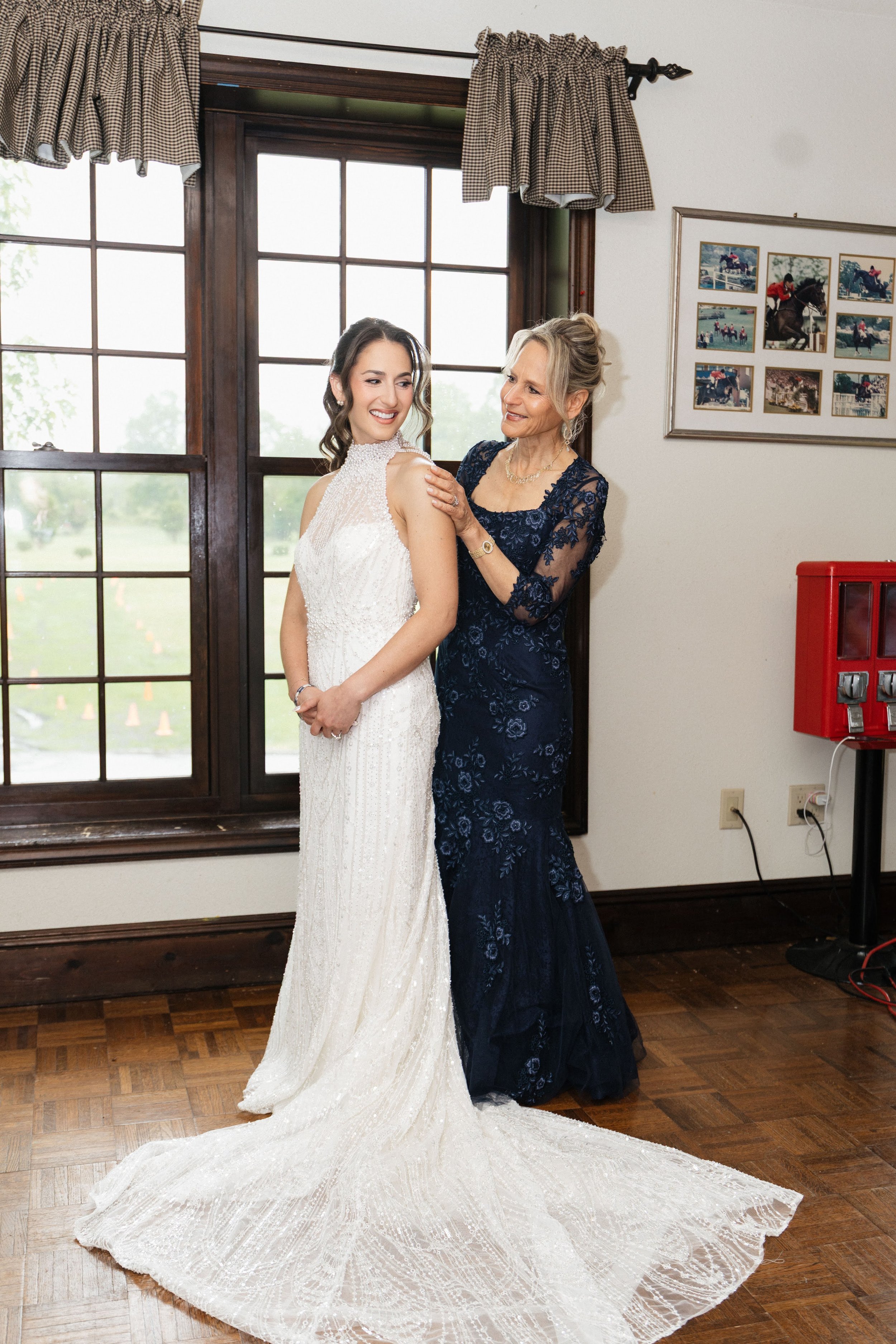 A bride in a white wedding dress and an older woman in a blue lace gown standing indoors near a window with checkered curtains. The bride is smiling while the older woman looks at her affectionately. There are framed photos of horse racing on the wal