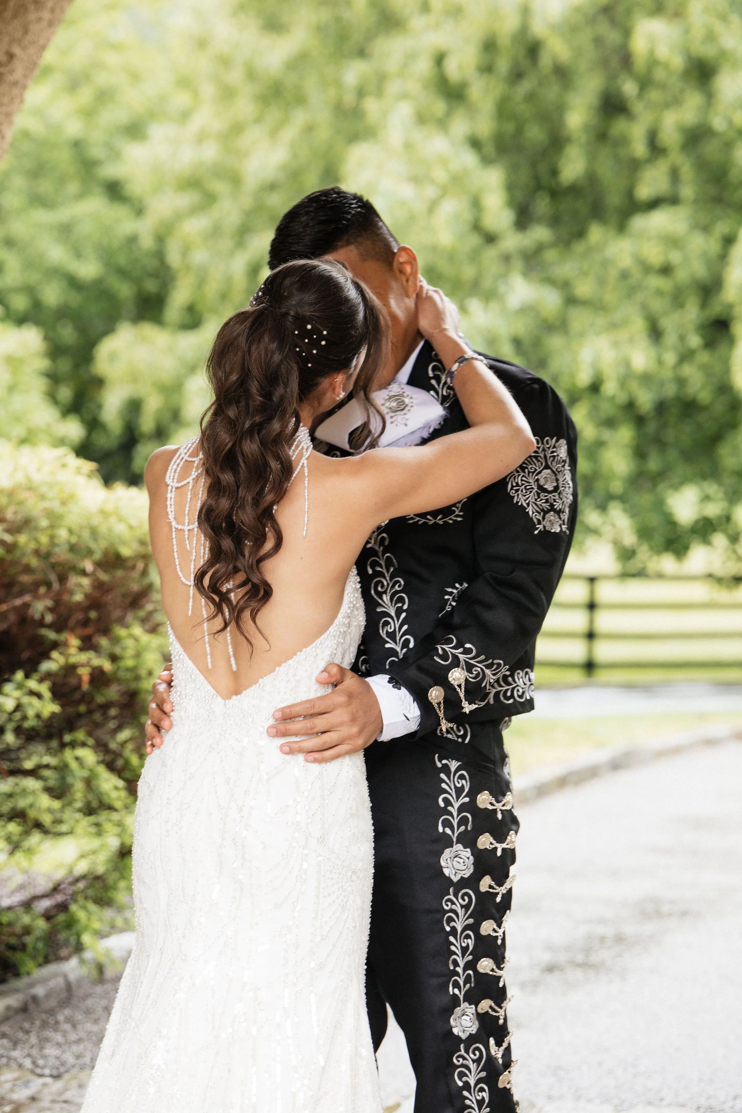 A bride and groom embracing and kissing outdoors, with trees and greenery in the background. The bride is wearing a white wedding dress with pearl details, and the groom is in a black embroidered suit.