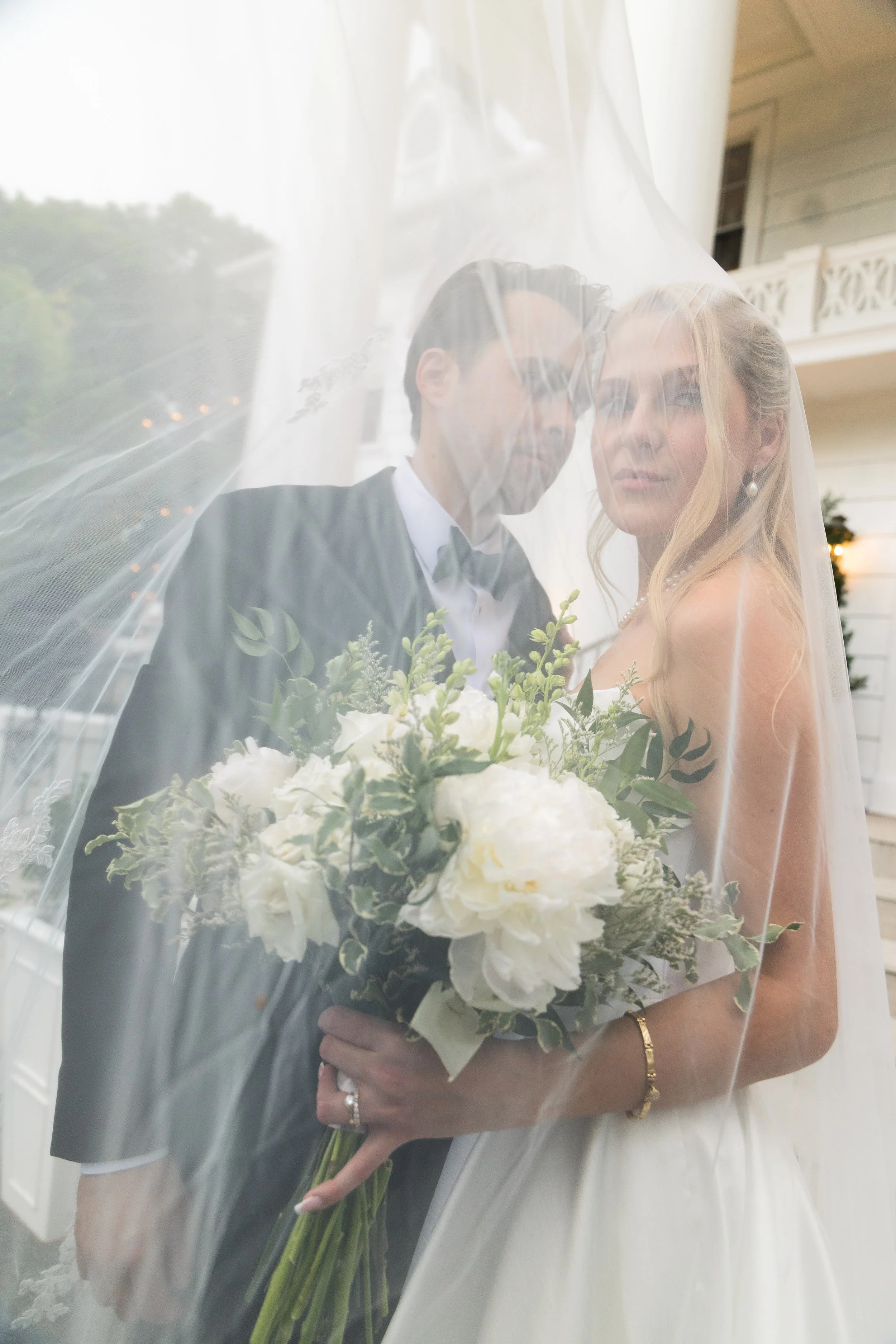 Bride and groom behind a veil, holding a bouquet of white flowers with greenery, during their wedding ceremony outside a house.