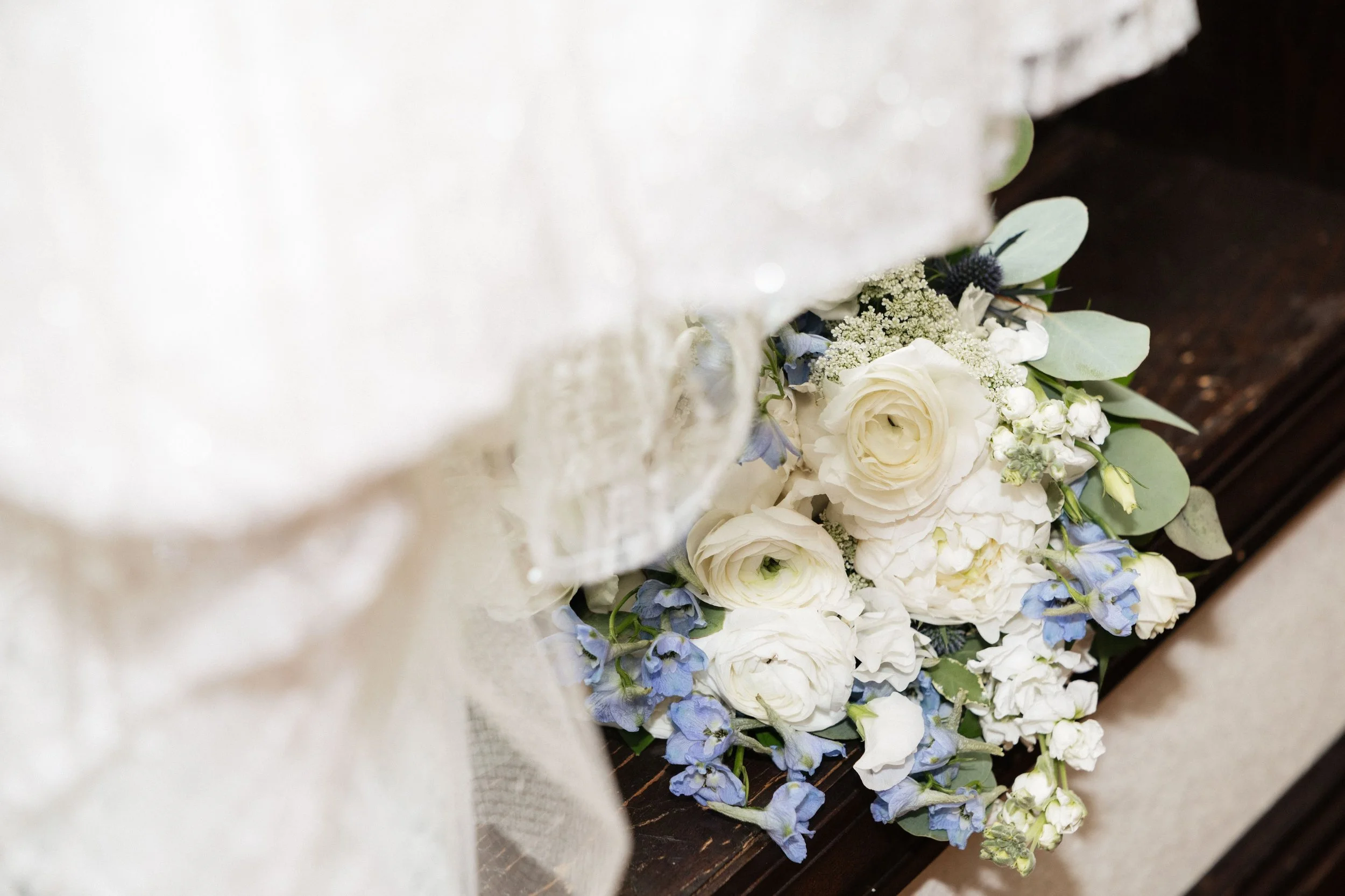 Close-up of a floral arrangement of white roses, blue and white flowers, and green leaves on a dark wooden surface.