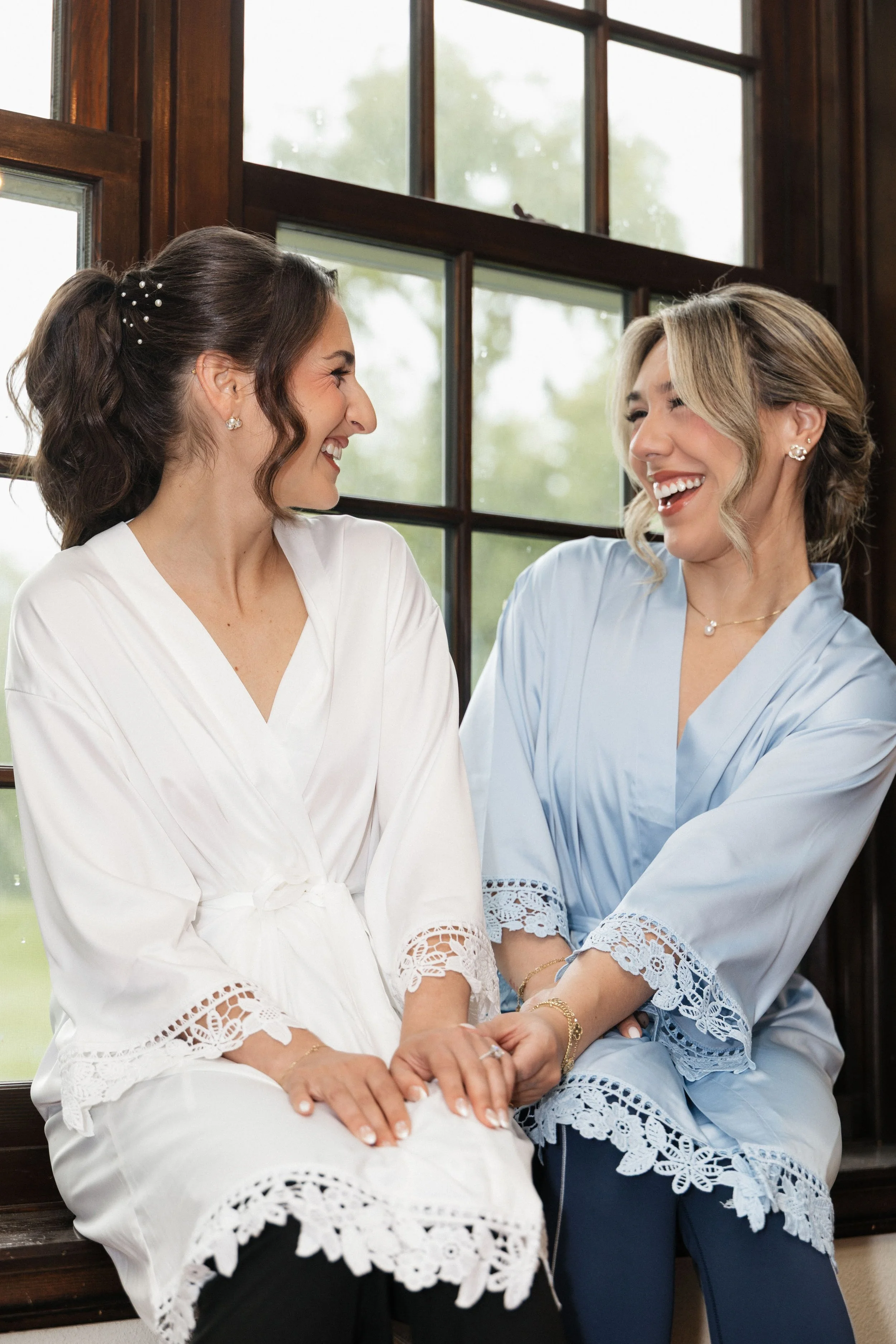 Two women sitting close together by a window, smiling, wearing matching pastel-colored robes with lace details, holding hands and looking happily at each other.
