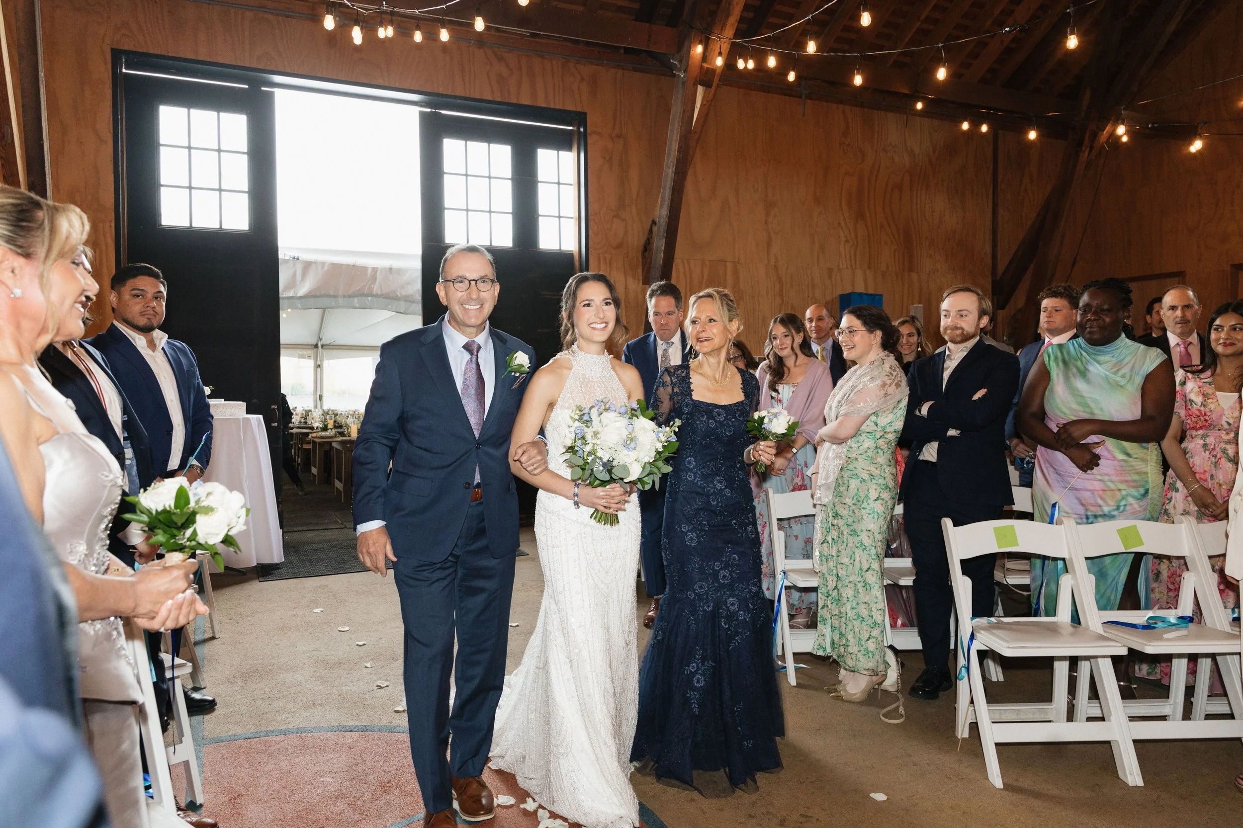 A bride and groom walking down the aisle at a wedding ceremony with guests standing on both sides, inside a rustic wooden venue decorated with string lights.