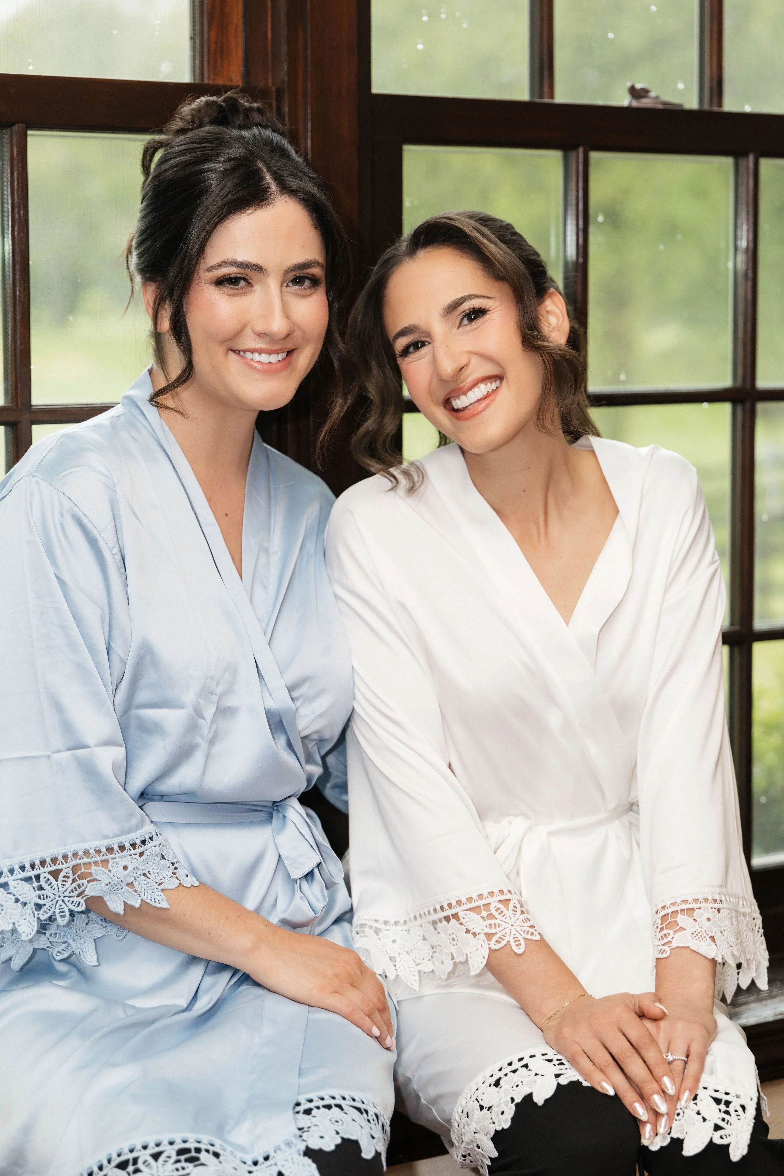 Two women sitting closely together, smiling near a window with outside greenery visible, wearing white and light blue dresses with lace details at the hem.