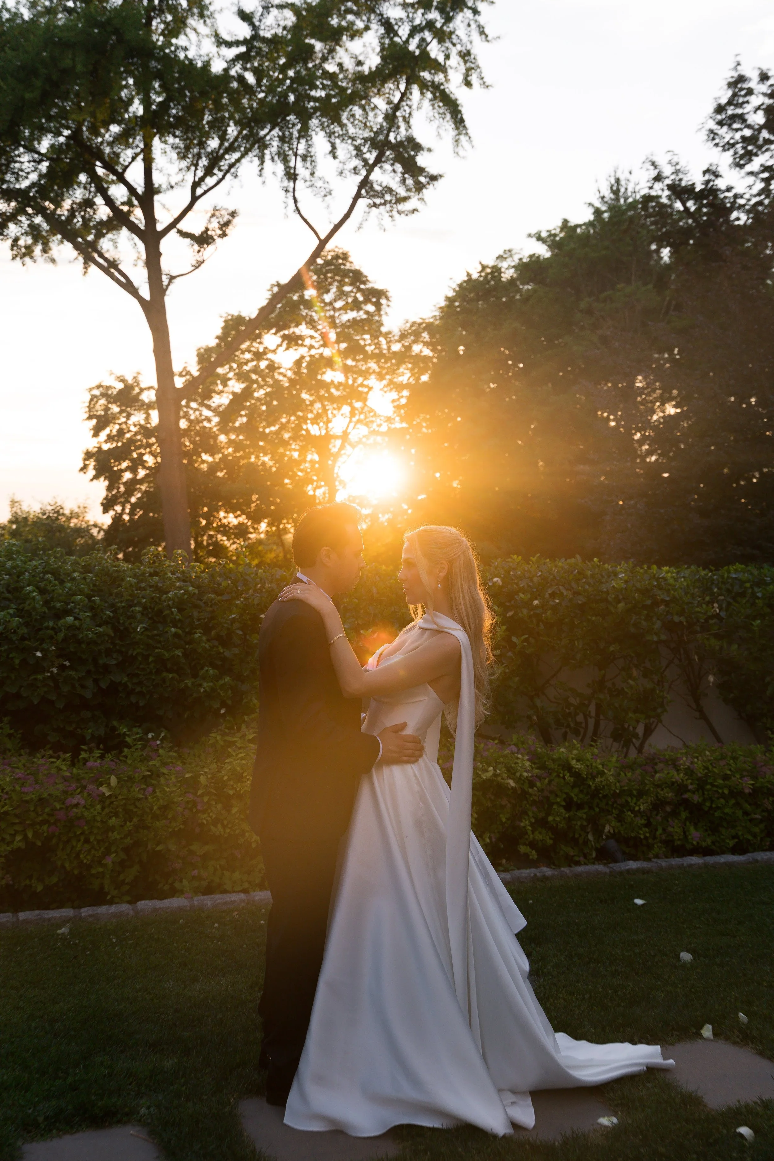 A bride and groom in wedding attire holding each other and facing each other outdoors during sunset, with trees and a hedge in the background.