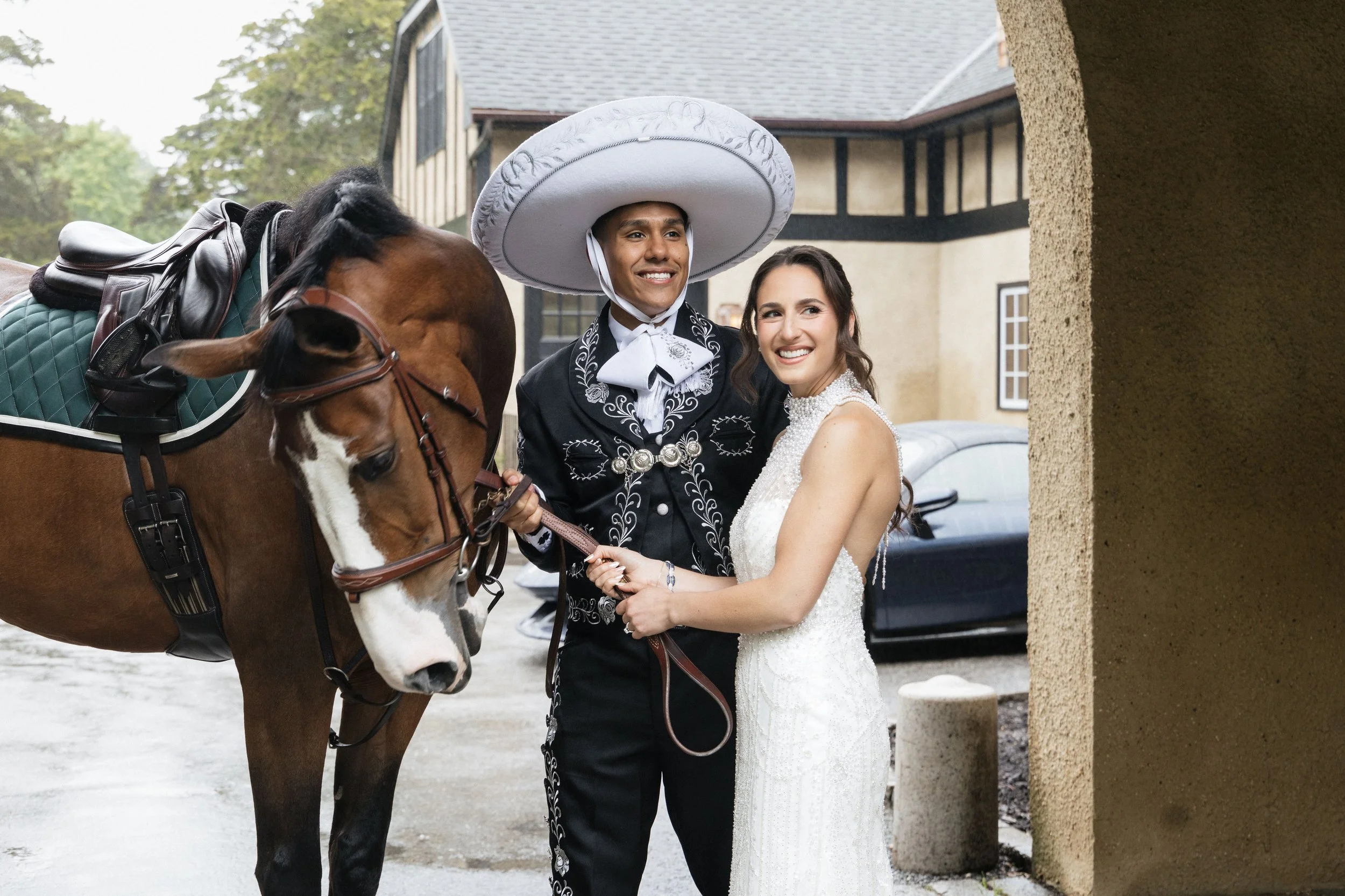 A smiling bride and groom in wedding attire standing by a horse with a bridle and saddle, under a stone archway in front of a building with beige walls and a gray roof, during a wedding photo shoot.