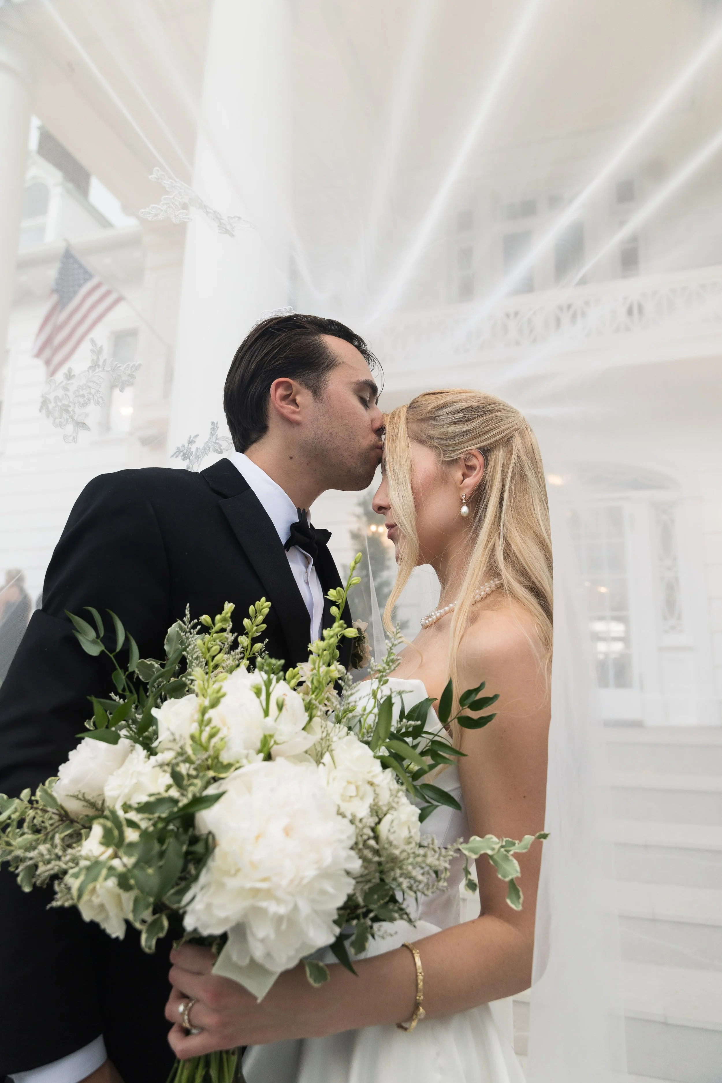 A bride and groom sharing a kiss at their wedding, with the bride holding a bouquet of white flowers and greenery, in an indoor venue with white walls and windows.