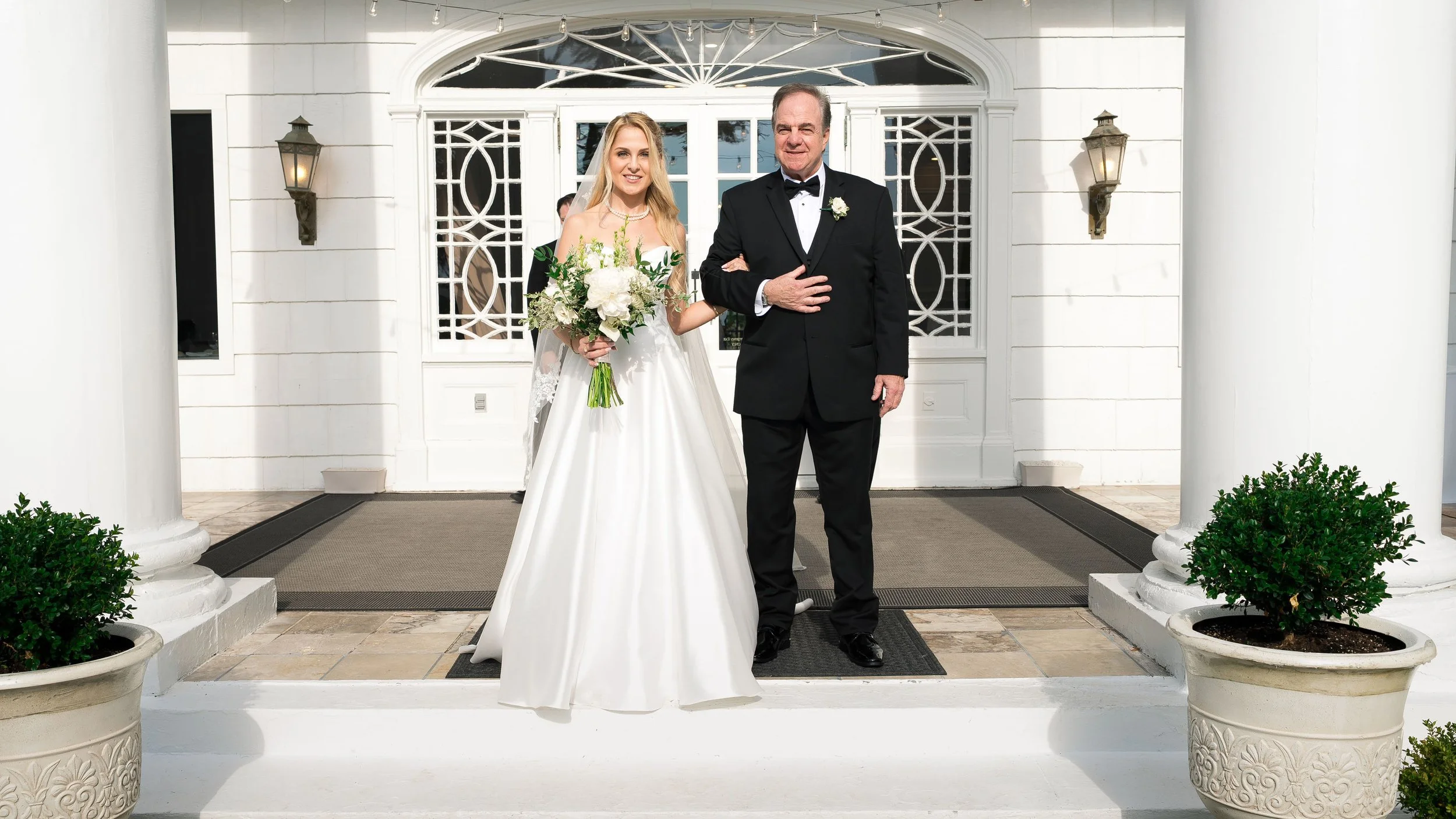 A bride in a white wedding gown holding a bouquet of white flowers and greenery, standing next to a man in a black tuxedo with a white shirt, black bow tie, and boutonniere, in front of a white building with decorative black window panels and two lan