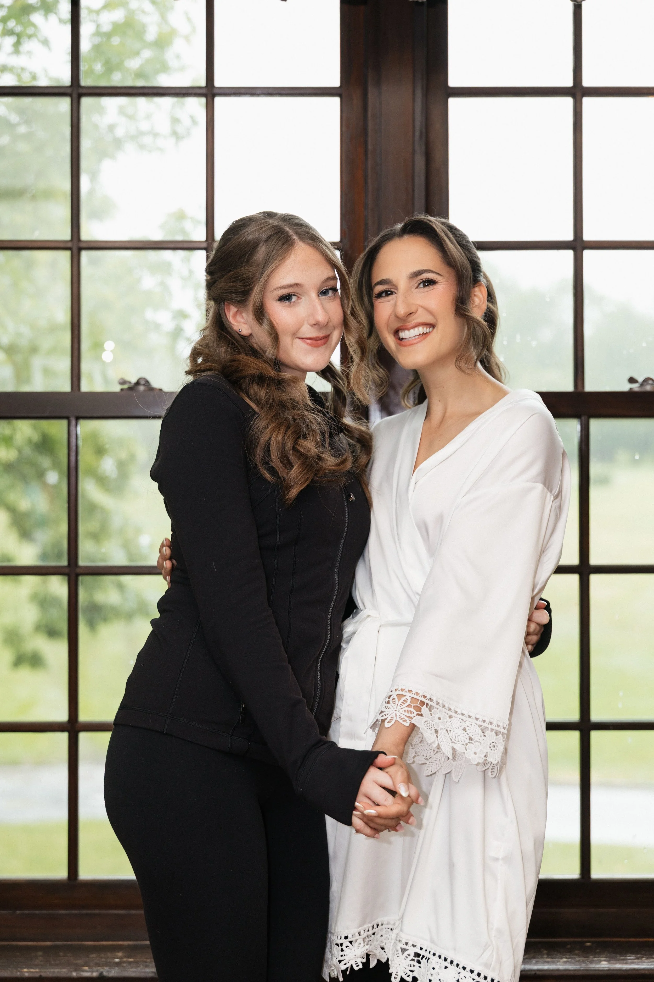 Two women stand close together, smiling and holding hands in front of large window panes with green trees outside. One woman wears a black jacket, and the other wears a white dress with lace details.
