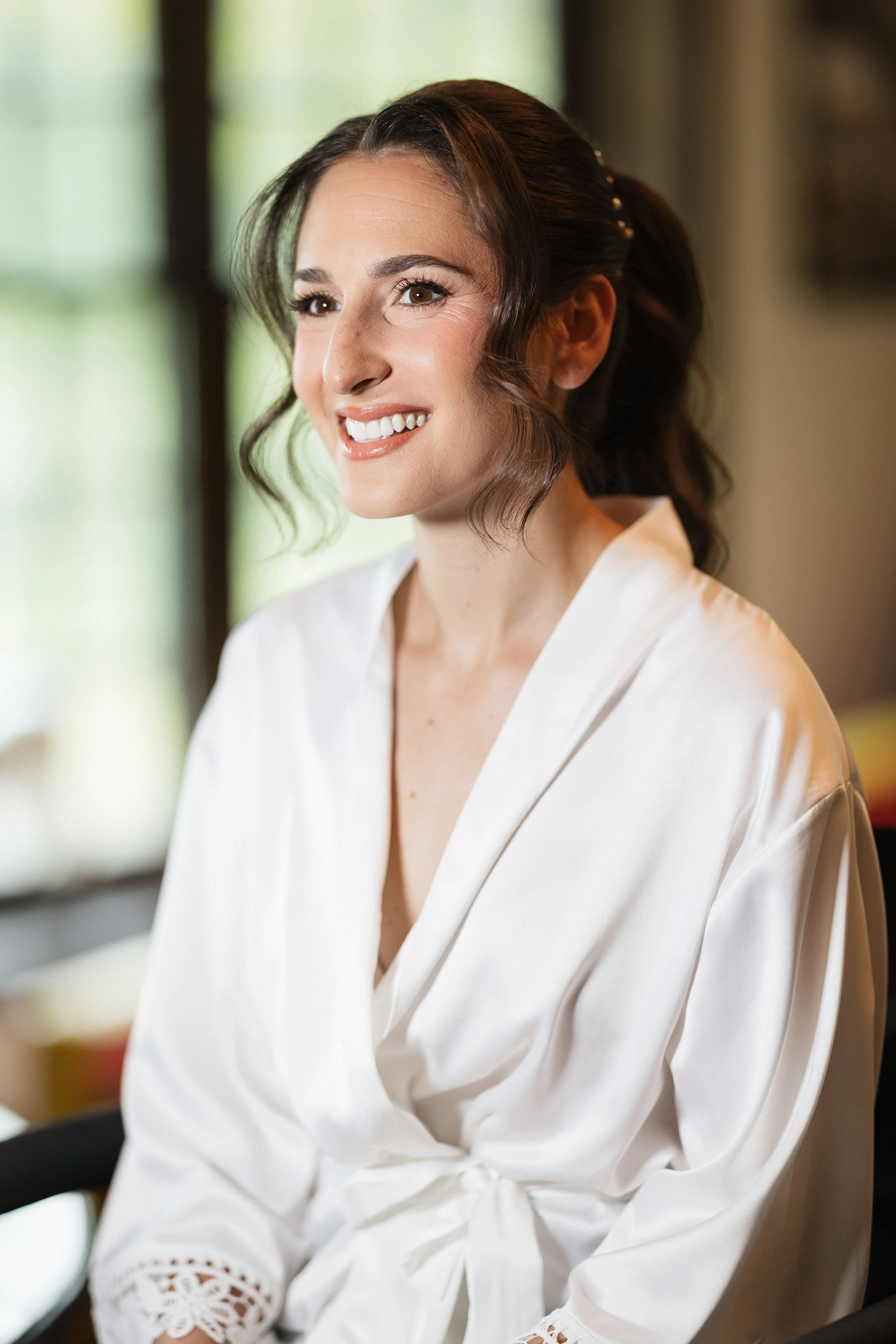 A woman with brown hair styled in loose waves, wearing a white satin robe with lace embroidery on the sleeves, smiling and looking to her right, indoors near a window with blurred greenery outside.