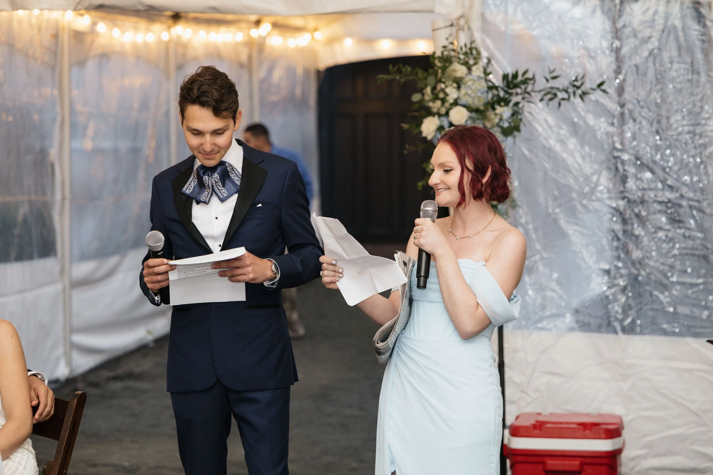 A young man in a dark tuxedo with a patterned bow tie and a young woman in a light blue dress reading and speaking at an indoor wedding reception with string lights and floral decorations.