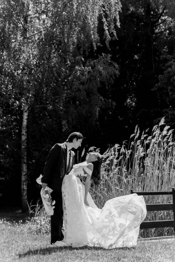 A black and white photo of a bride in a wedding dress sitting on a bench outdoors, with a person in a suit leaning over her, possibly adjusting her dress or veil. They are surrounded by tall grass and trees in the background.