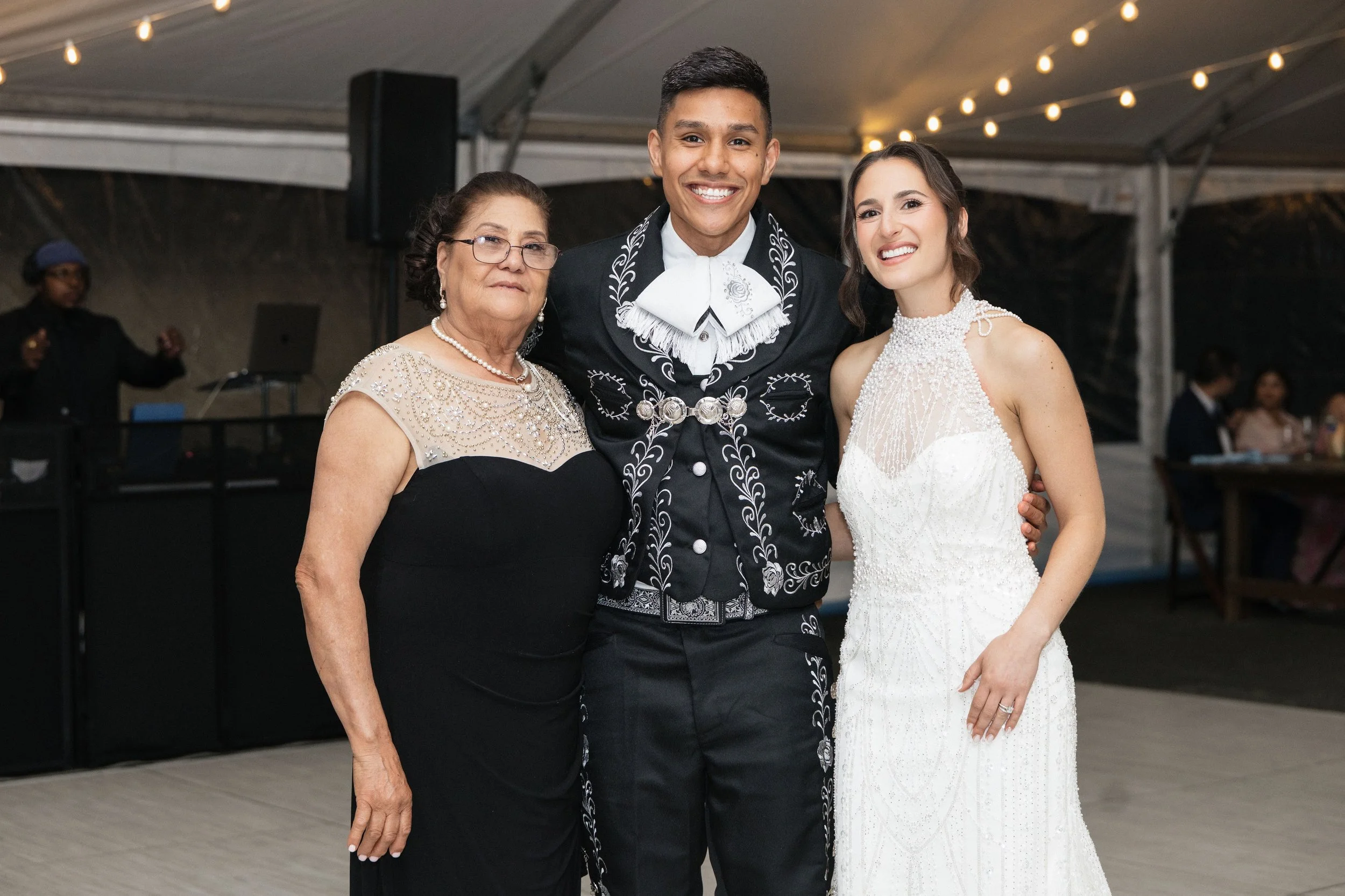 A young man in a bolo tie and traditional attire standing between an older woman in glasses and a young woman in a white dress, all smiling at a wedding celebration under a tent with string lights.